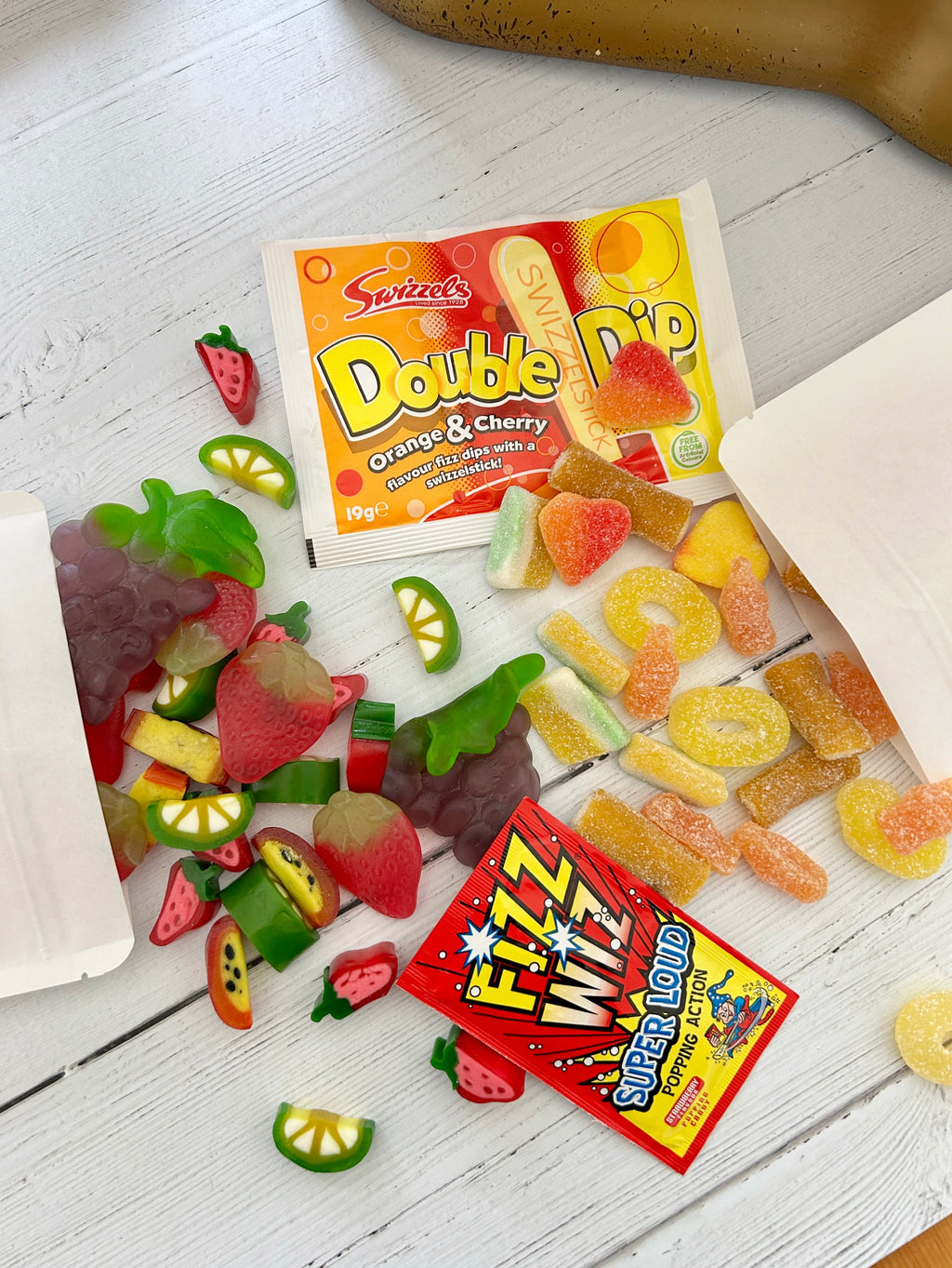 Summery Pick 'n' Mix sweets spilling out of white pouch packaging, double dip and fizz wiz sweets on white wooden surface.