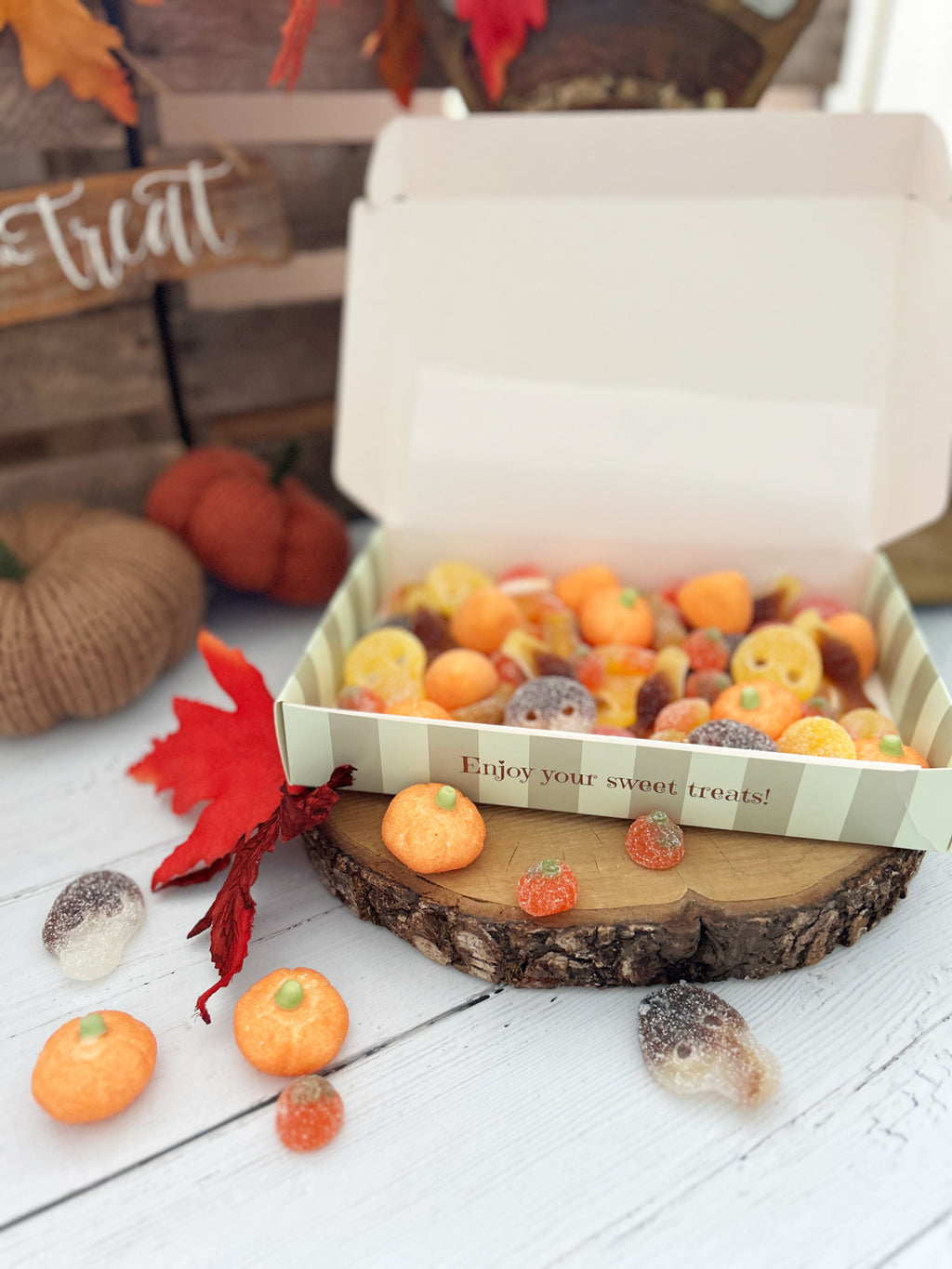 An open white gift box filled with assorted Halloween-themed candy treats, styled on a wood slice with small orange pumpkins, a red leaf, and autumn decorations in the background.