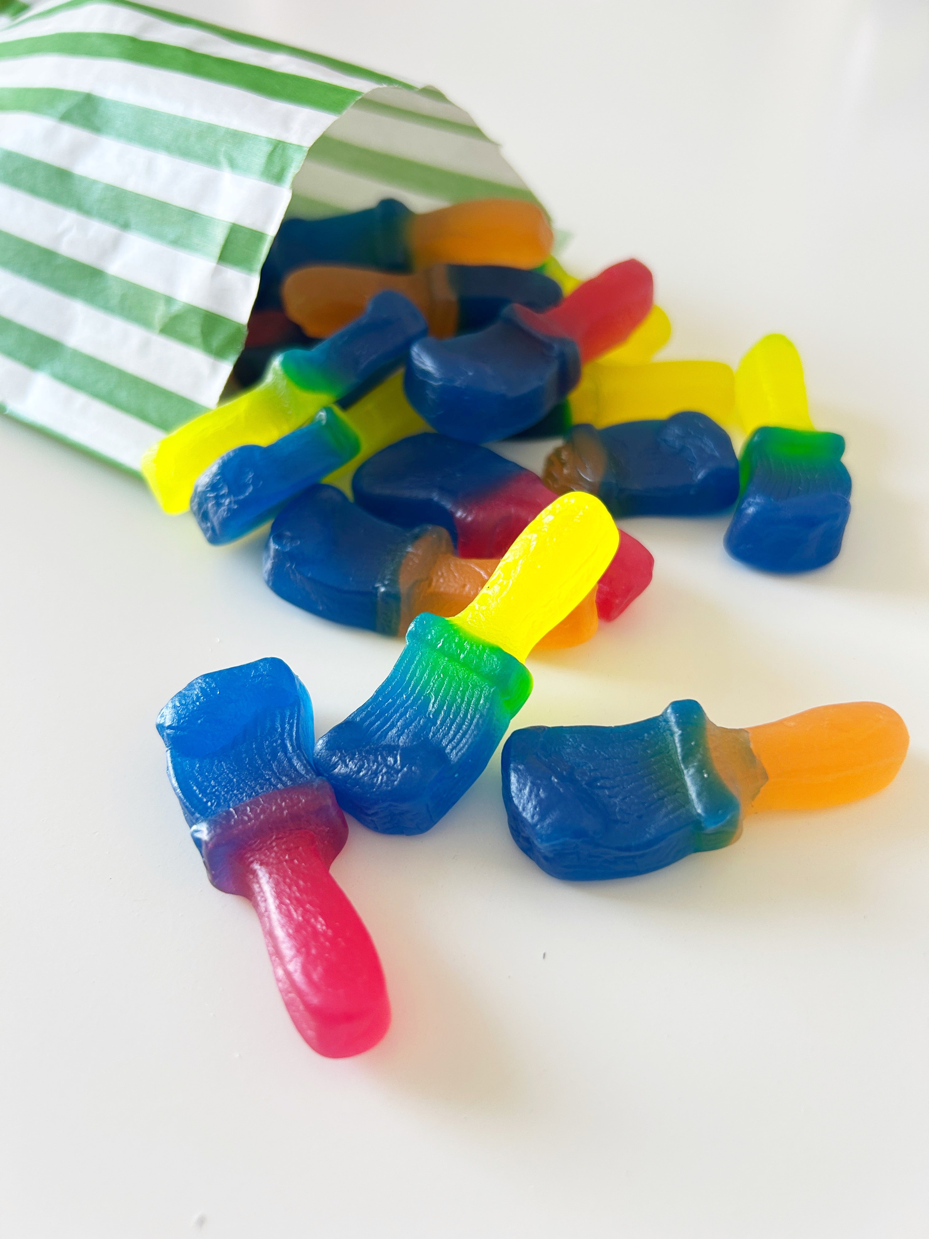 Multicolored gummy candies shaped like paint brushes spilling out of a green and white striped paper bag onto a white surface.