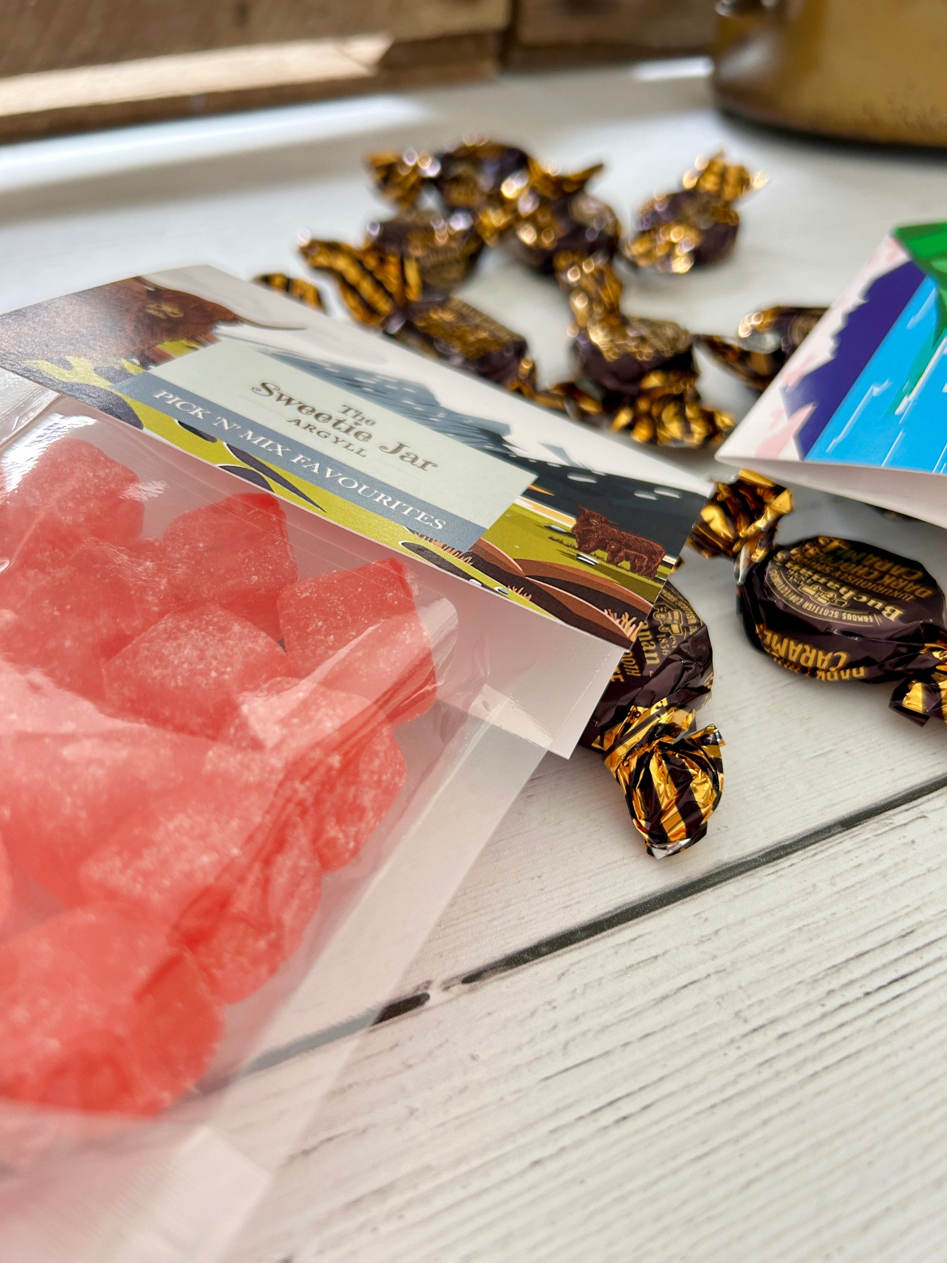 Two clear bags of Sweetie Jar sweets and chocolates arranged on a white wooden surface, with several individually wrapped dark chocolates scattered around them. One bag contains bright pink/red boiled candies and the other contains assorted colourful candies.