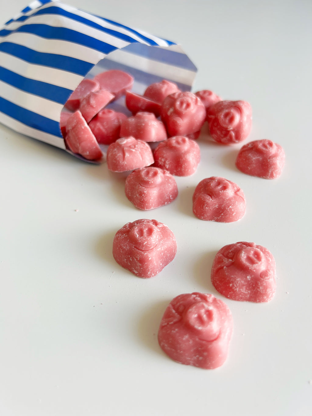Pink, pig shaped sweets spilling out of a blue and white striped paper bag onto a clean white surface.