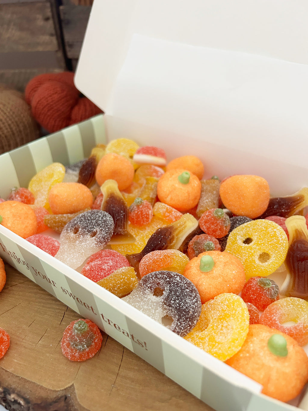 A white gift box filled with assorted colorful gummy candies, including orange pumpkin-shaped gummies, sugar-coated ring gummies, and ghost gummies, set on a wooden surface.
