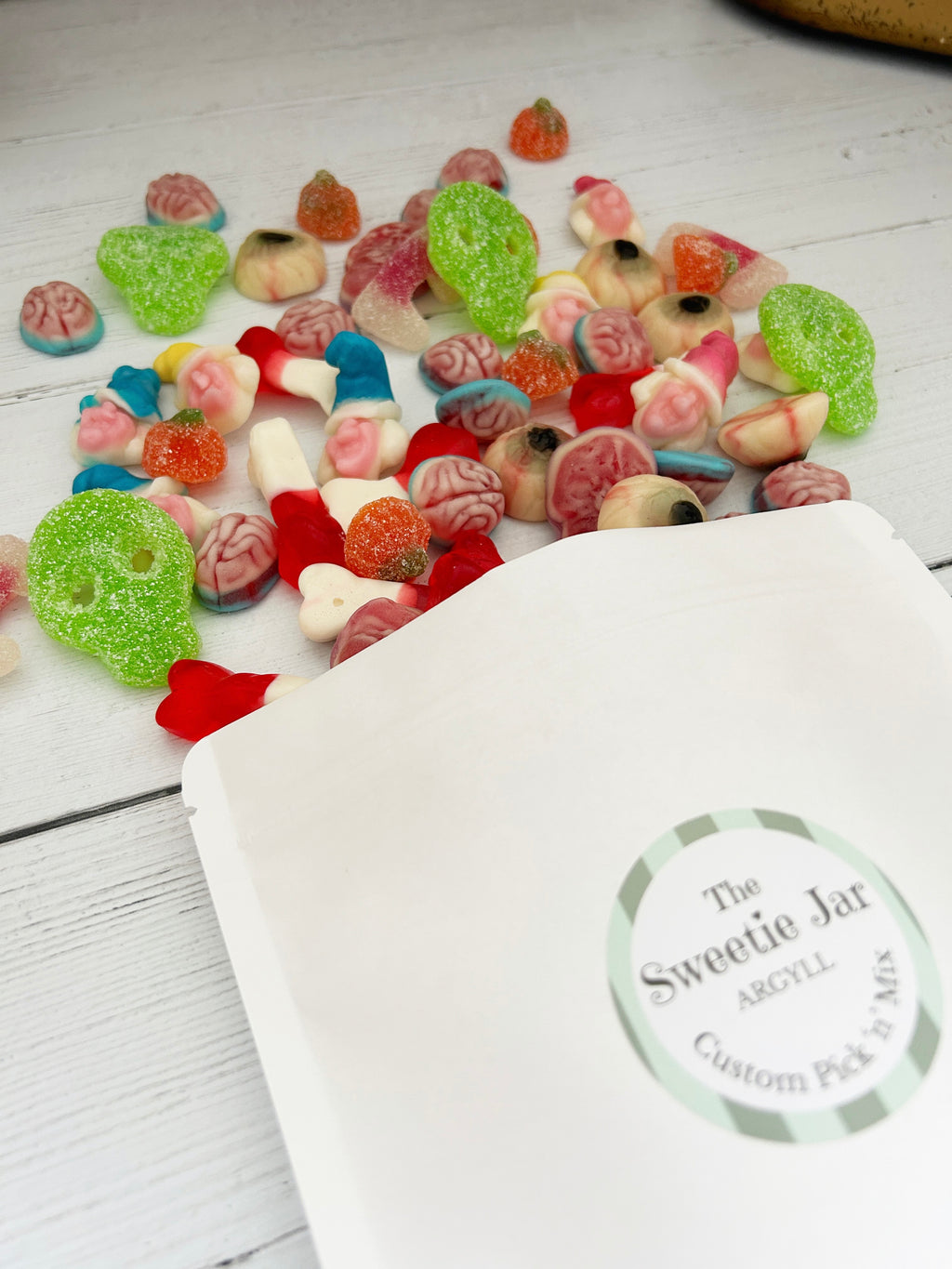 A white stand-up pouch labeled 'The Sweetie Jar' with 'Custom Pick & Mix' printed on the front, shown on a white wood surface with assorted colorful gummy candies spilled out above it.