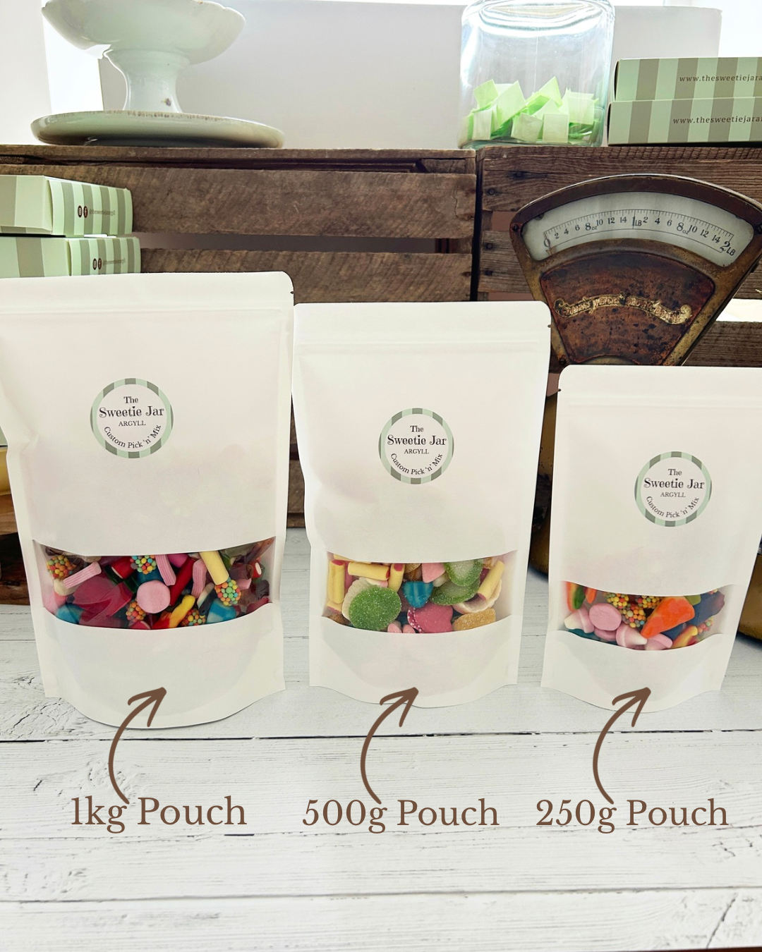 Three white stand-up pouches of 'The Sweetie Jar' displayed on a white wooden surface, with size labels indicating 1kg, 500g, and 250g. The clear lower window of each pouch reveals colourful pick n mix sweets inside.