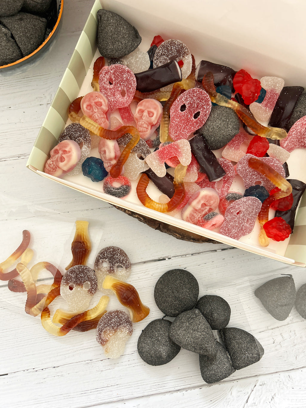A box of assorted Halloween-themed gummy candies and sugar-coated treats, including ghost gummies, dark chocolate pieces, and gray stone-like candies, arranged on a white wooden surface.