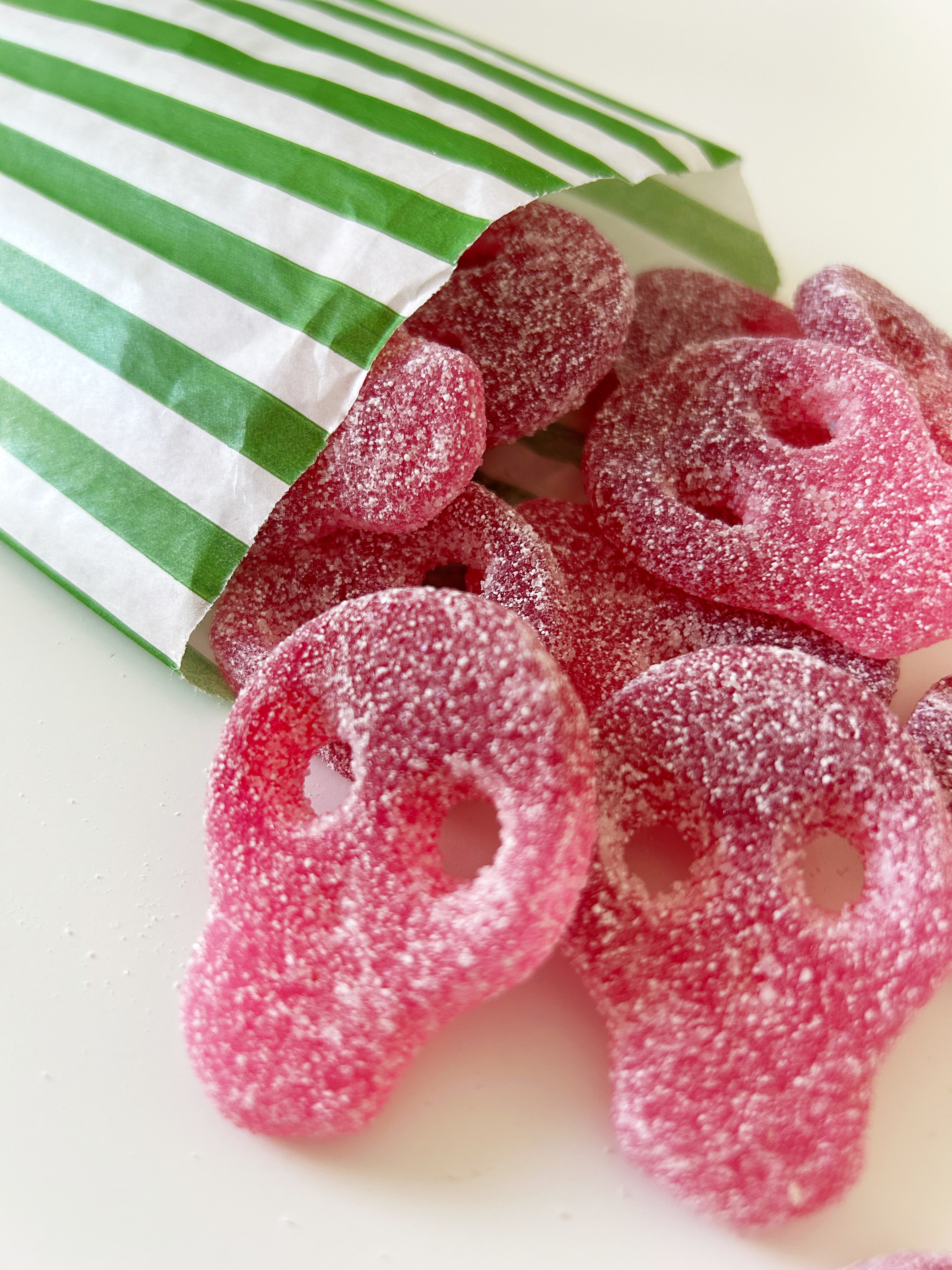 Pink skull-shaped gummy candies coated in sugar, spilling out of a green and white striped paper bag on a white surface.