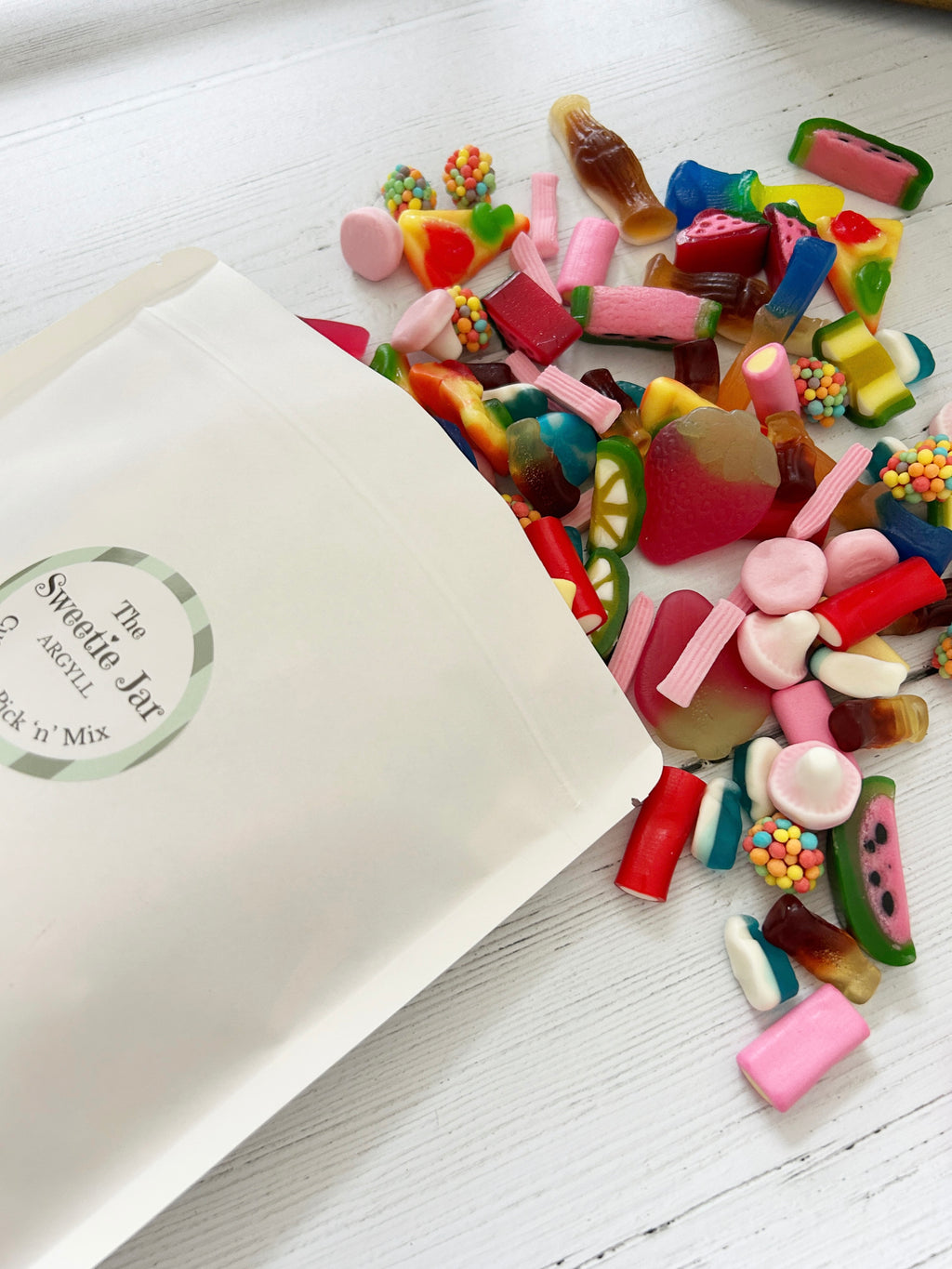 The Sweetie Jar custom pick 'n' mix pouch with sweets spilling out of it onto white wooden surface.