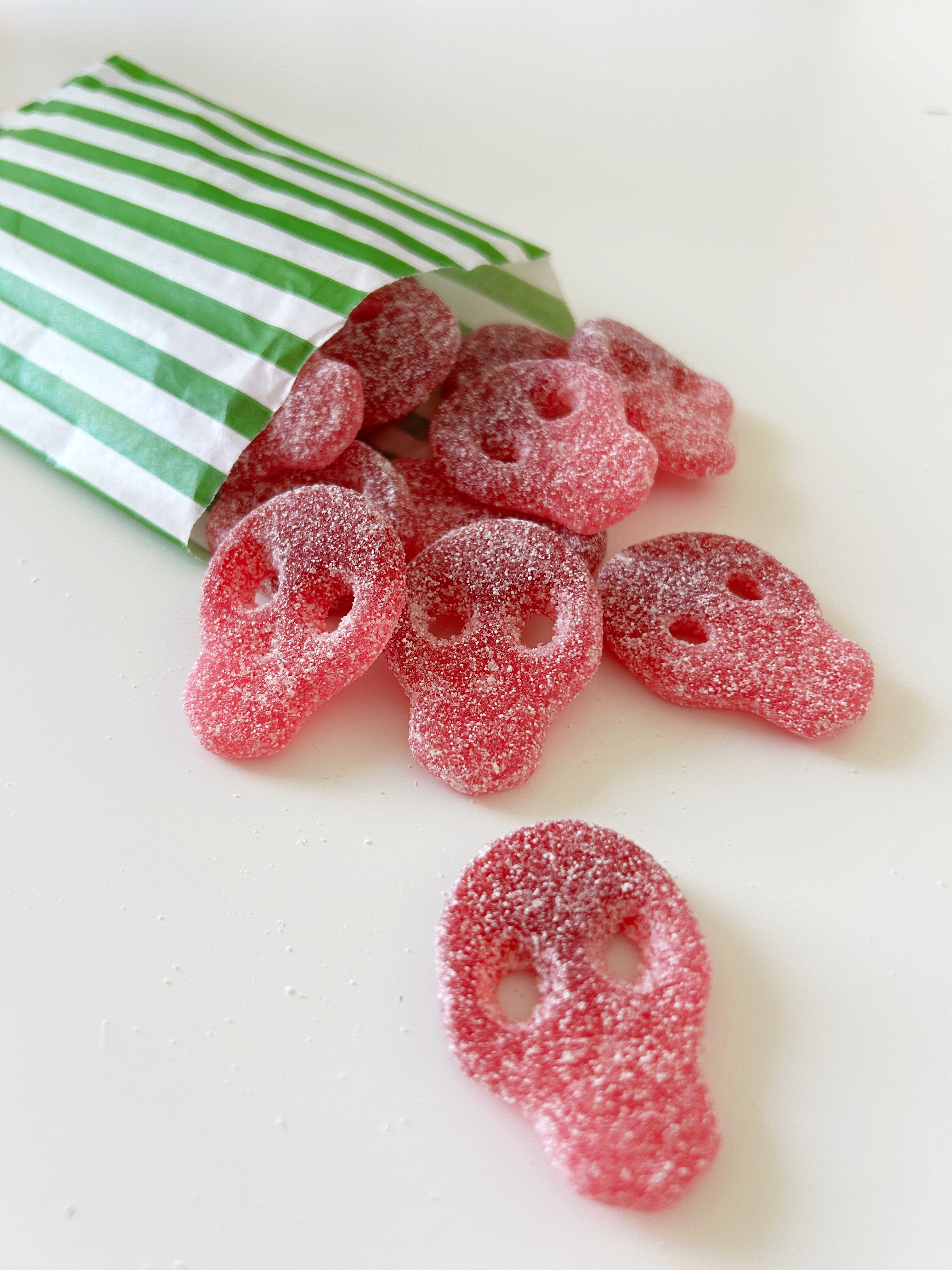 Pink sugar-coated skull-shaped gummy candies spilling from a green and white striped paper bag on a white surface.