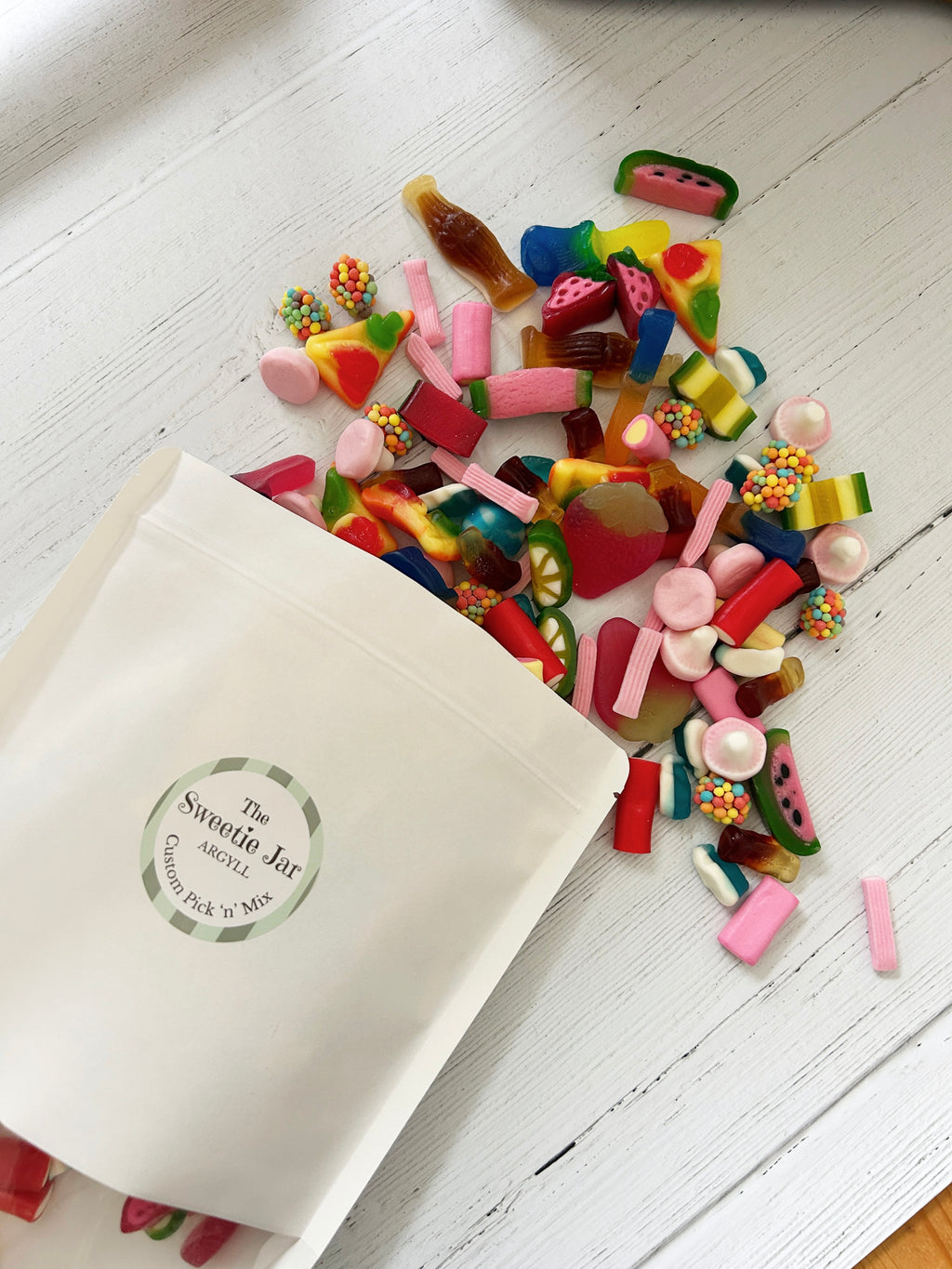 The Sweetie Jar custom pick 'n' mix pouch with sweets spilling out of it onto white wooden surface.