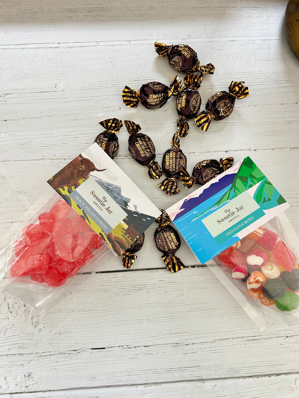 Two clear bags of Sweetie Jar sweets and chocolates arranged on a white wooden surface, with several individually wrapped dark chocolates scattered around them. One bag contains bright pink/red boiled candies and the other contains assorted colourful candies.