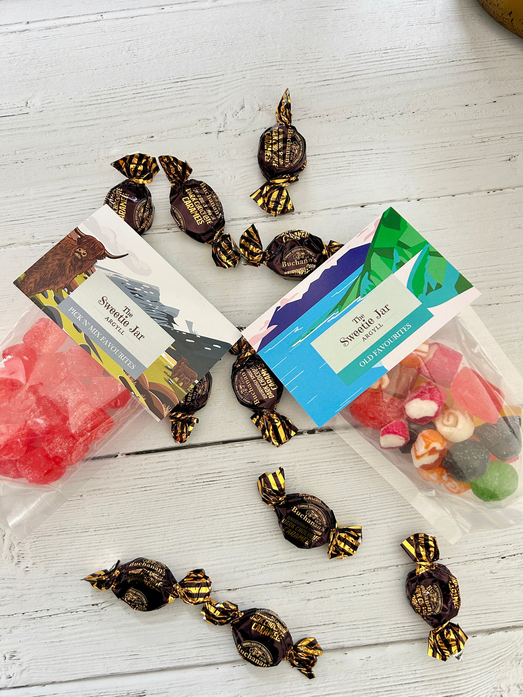 Two clear bags of Sweetie Jar sweets and chocolates arranged on a white wooden surface, with several individually wrapped dark chocolates scattered around them. One bag contains bright pink/red boiled candies and the other contains assorted colourful candies.