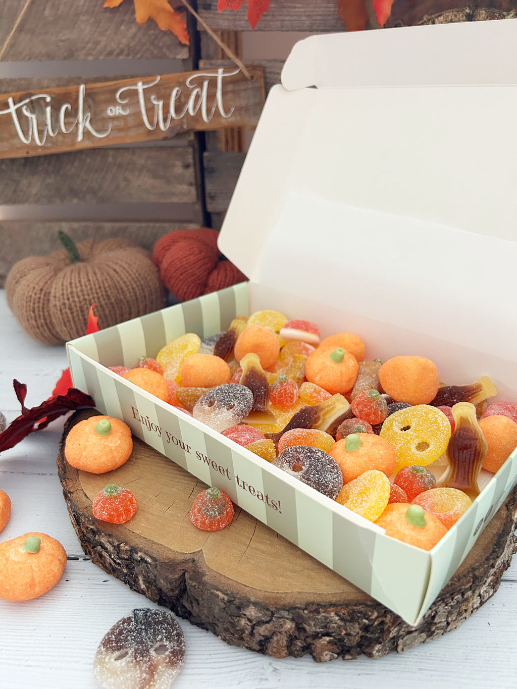 An open white gift box filled with assorted Halloween-themed gummy candies, including orange pumpkins, citrus slices, and sour candies, displayed on a rustic wood slice with fall decorations in the background.
