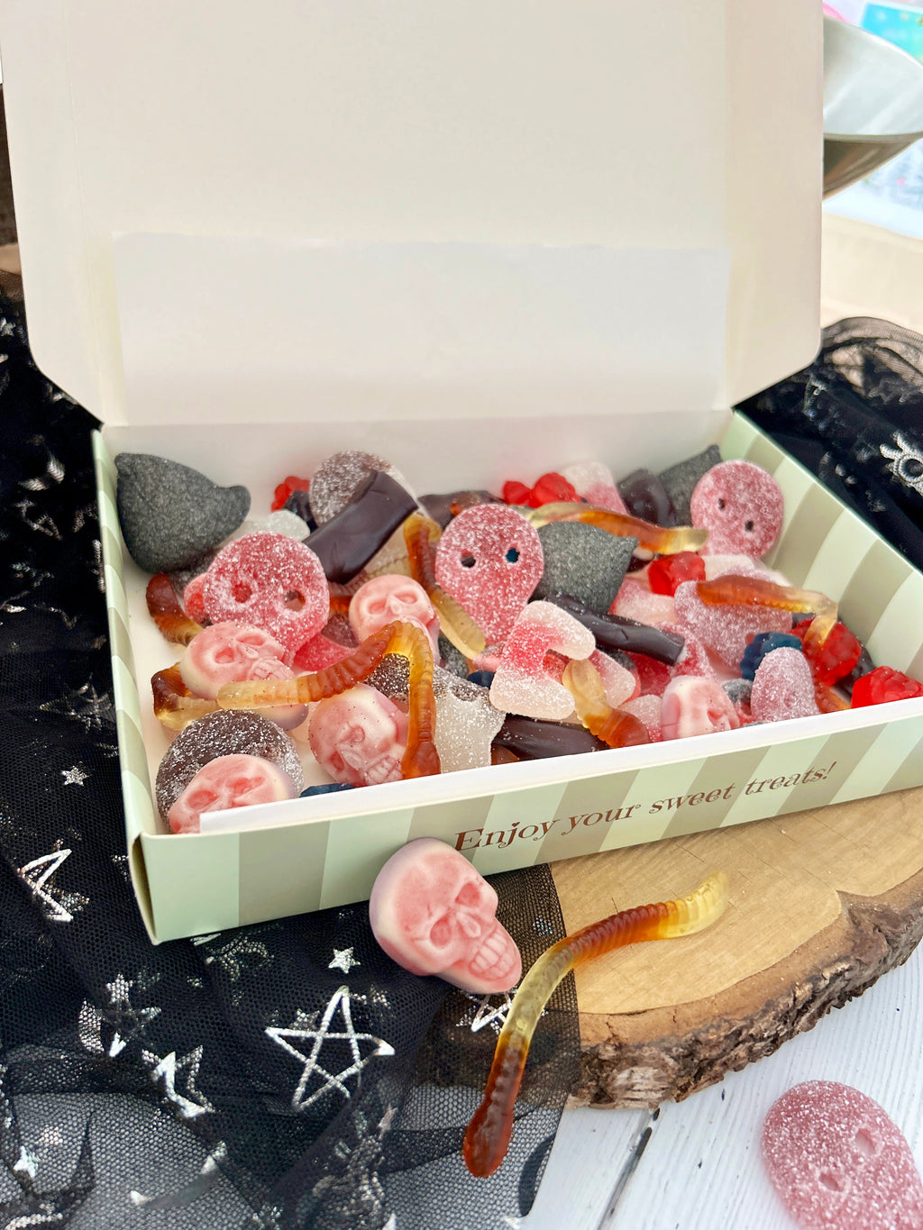 An open white gift box filled with assorted gummy candies and sour gummy worms, styled on a rustic wood slice with black tulle fabric featuring white star and pentagram patterns.