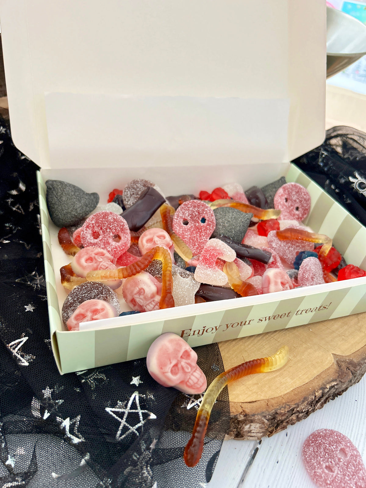 An open white gift box filled with assorted gummy candies and sour gummy worms, styled on a rustic wood slice with black tulle fabric featuring white star and pentagram patterns.