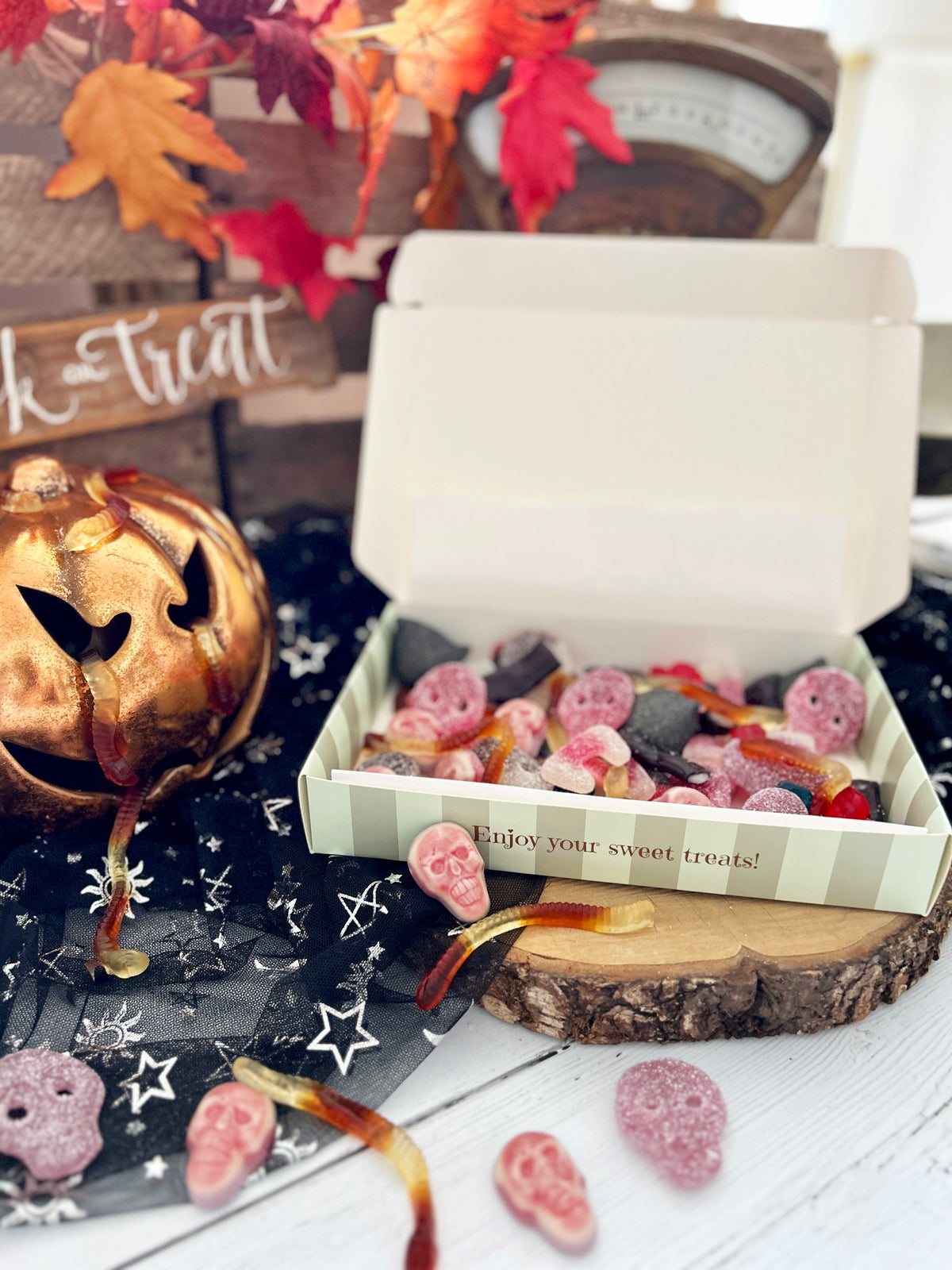 An open white gift box filled with assorted Halloween gummy and hard candies, styled on a rustic wood slice with scattered gummies and candy hearts. The scene includes a gold jack-o'-lantern, black spiderweb fabric, autumn leaves, and a sign reading 'Treat'.