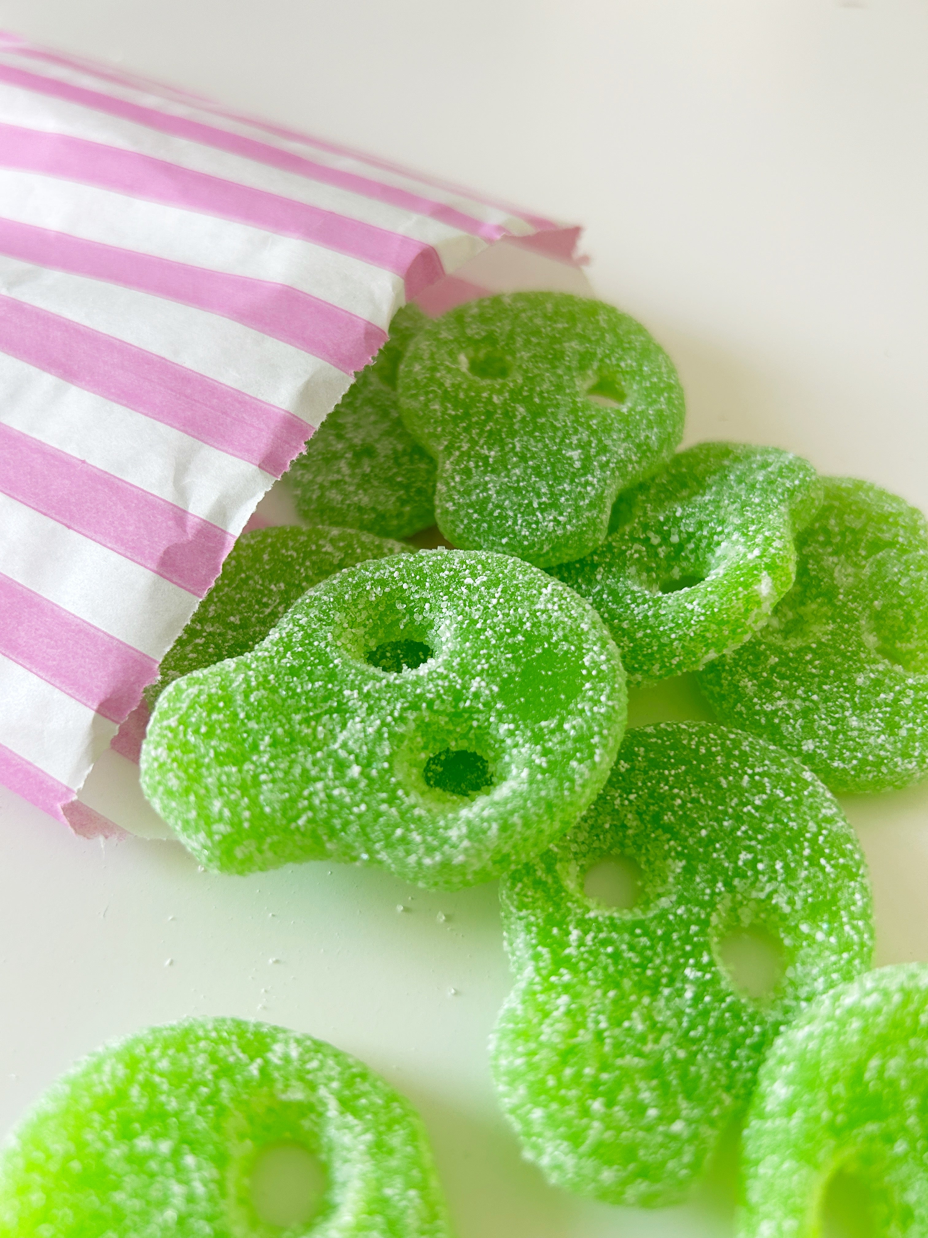 Green skull-shaped sour gummy candies coated in sugar, spilling from a pink and white striped paper bag on a white surface.