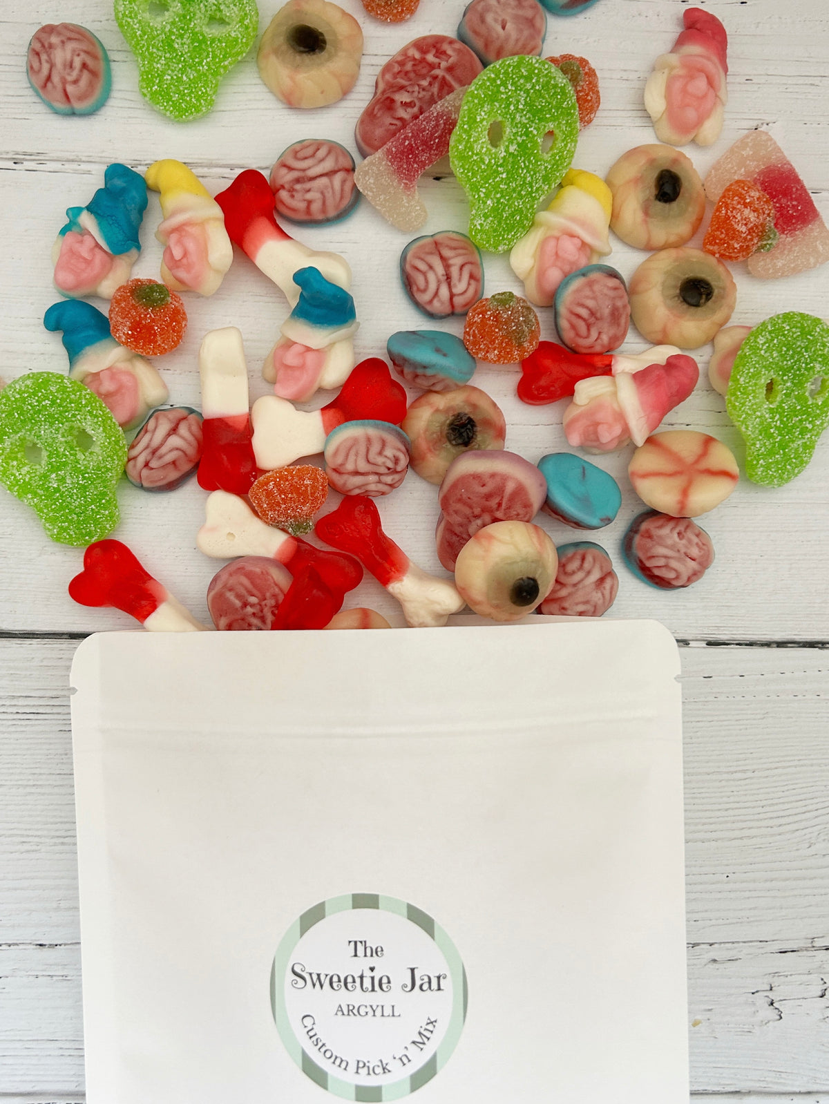 A white stand-up pouch labelled 'The Sweetie Jar Argyll Custom Pick 'N Mix' sits on a white wood surface below a scattered assortment of colourful novelty gummy candies, including brain-shaped, eye-shaped, and striped gummies.