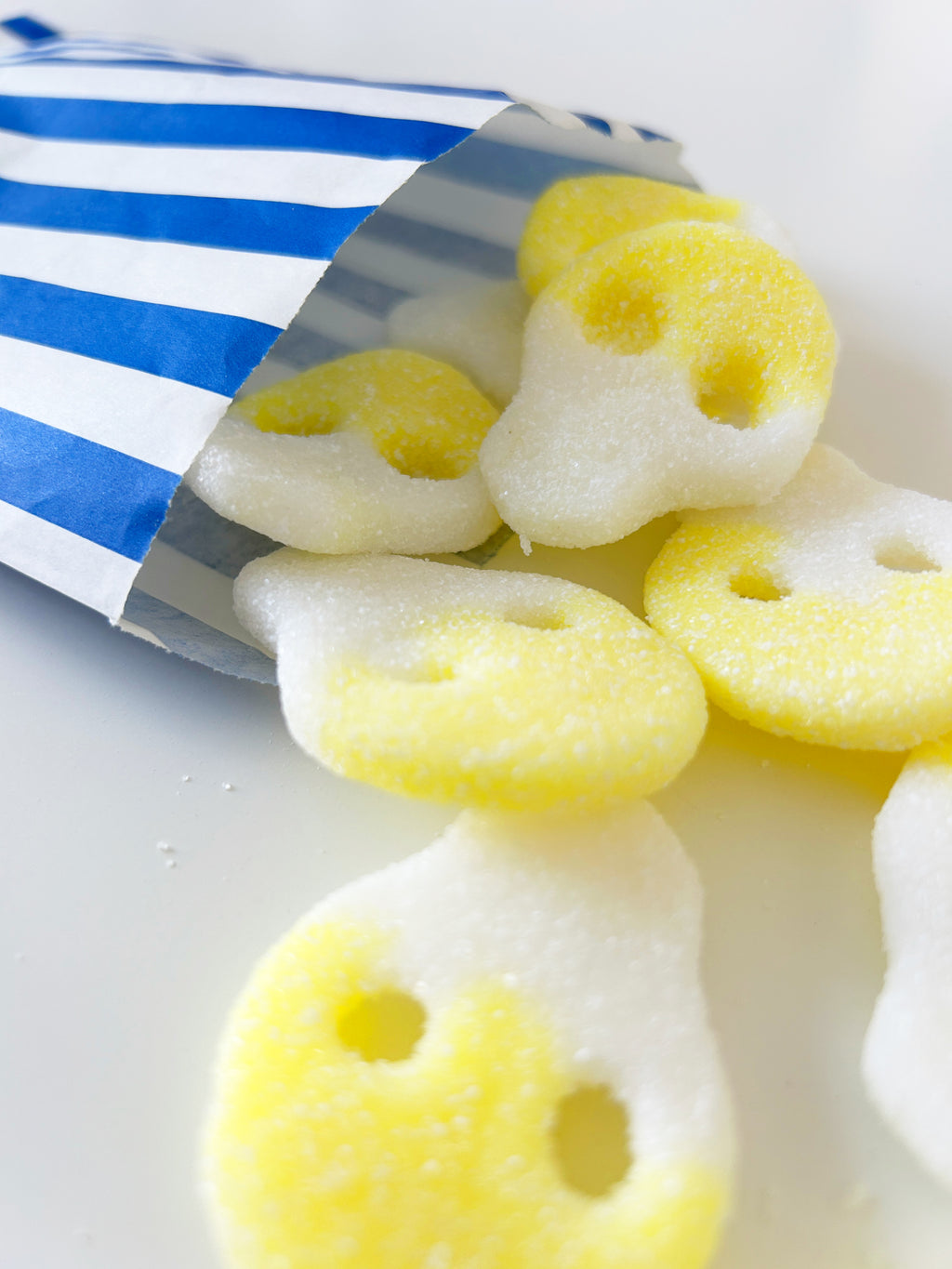 Yellow and white sugar-coated ghost-shaped gummy candies spilling out of a blue and white striped paper bag on a white surface.