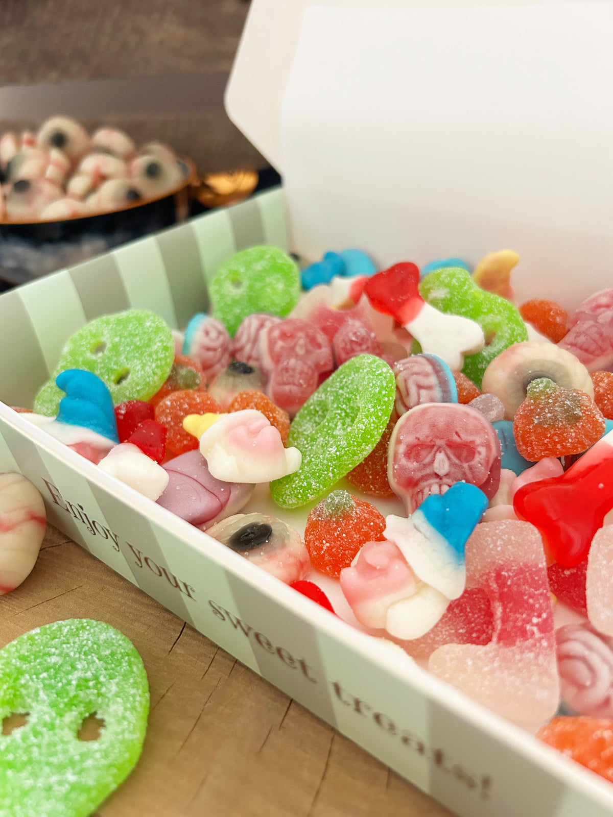 A white box filled with assorted colorful gummy candies, including skull-shaped gummies, green sugar-coated gummies, and pink and red gummies,.