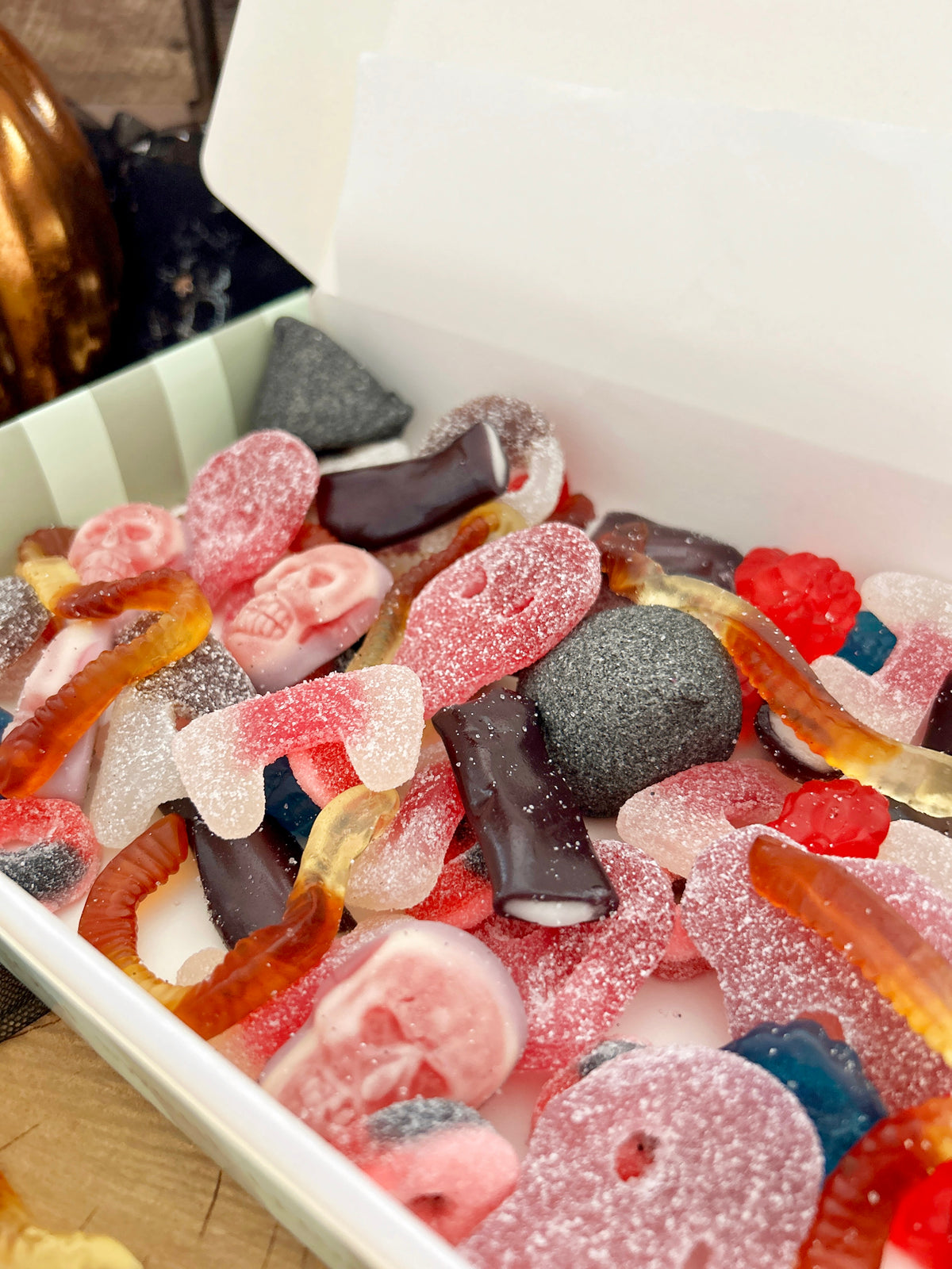 A close-up view of assorted gummy and soft candy pieces inside a white box, including pink, red, orange, brown, and dark gray pieces with sugar coating.