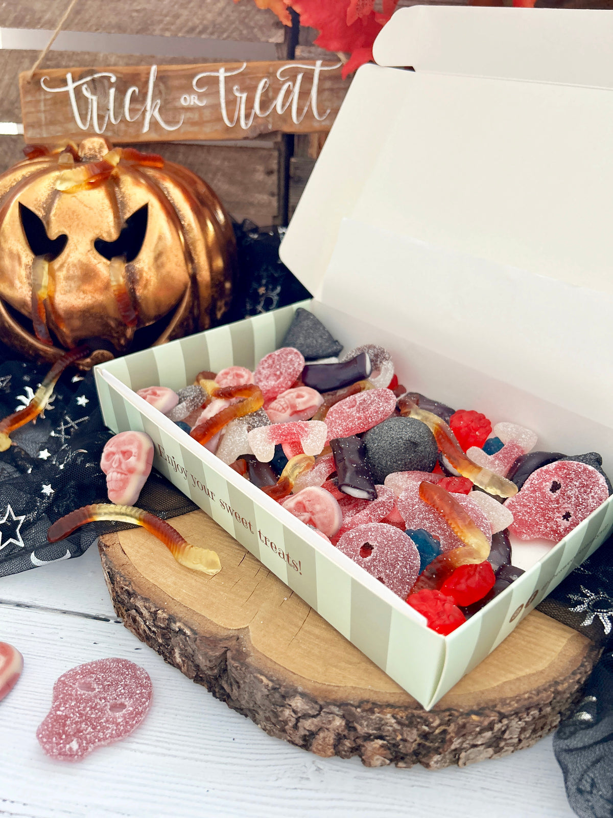 A Halloween-themed candy box filled with assorted gummy candies, shown open on a rustic wood slice with a carved pumpkin and fall decorations in the background. The box has a pale green and white striped exterior and the words 'trick or treat' appear above.