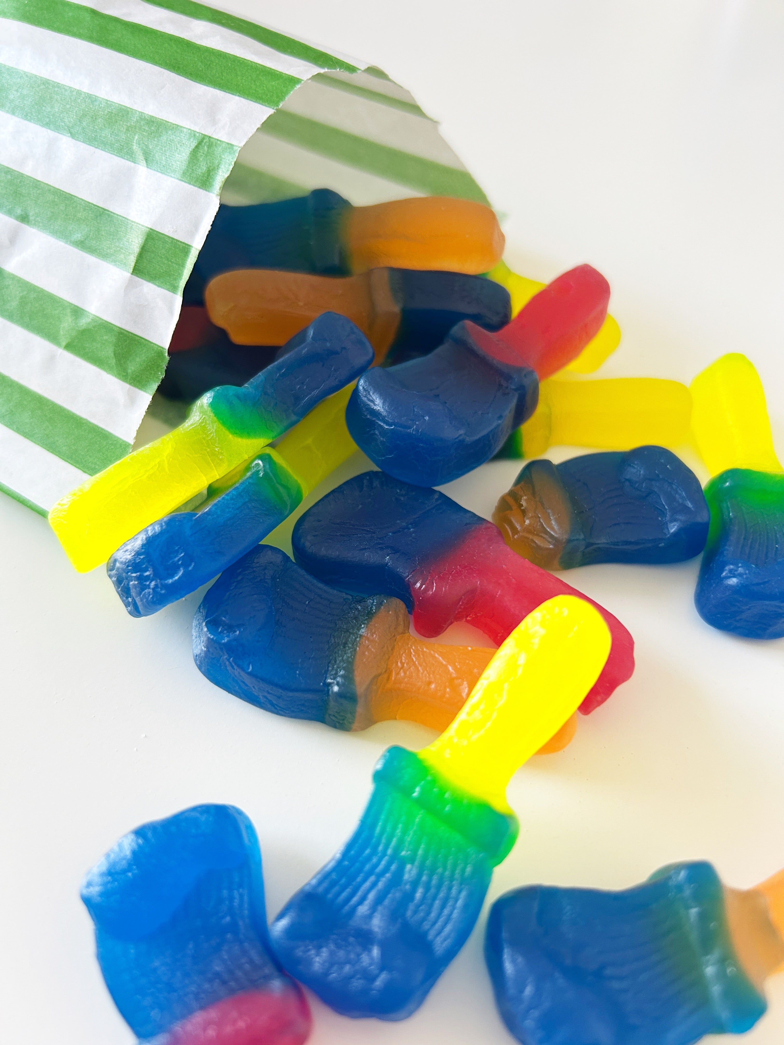Multicolored gummy sweets shaped like paintbrushes spilling out of a green and white striped paper bag onto a white surface.
