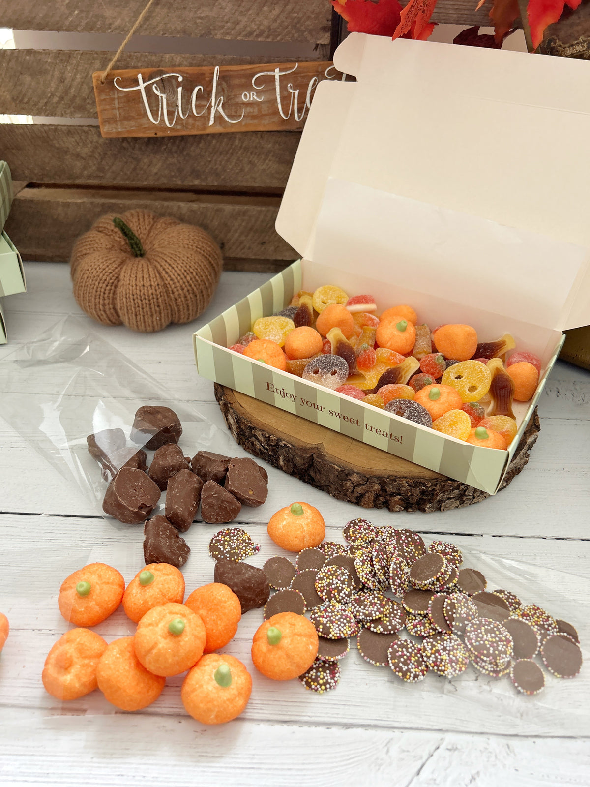 An open gift box filled with assorted fall-themed candies and chocolates sits on a rustic wood slice, with extra pumpkin-shaped candies and chocolate-covered treats scattered around on a white tabletop. A small knitted pumpkin and a wooden sign reading 'trick or treat' are in the background.