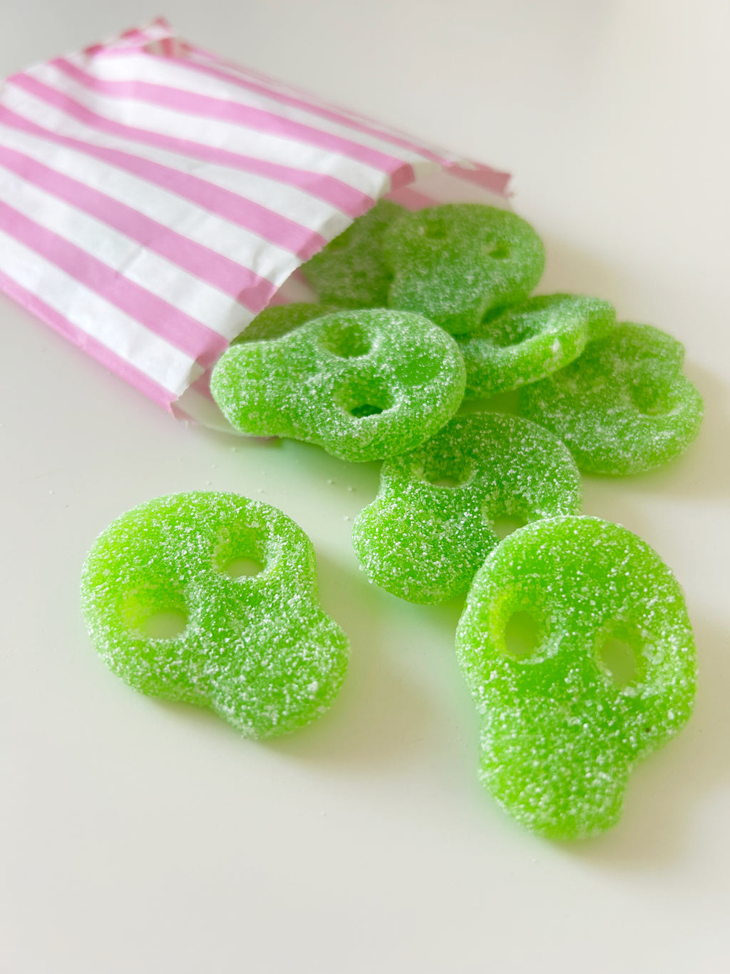 Green sugar skull gummy candies spilling out of a pink and white striped paper bag on a light cream background.