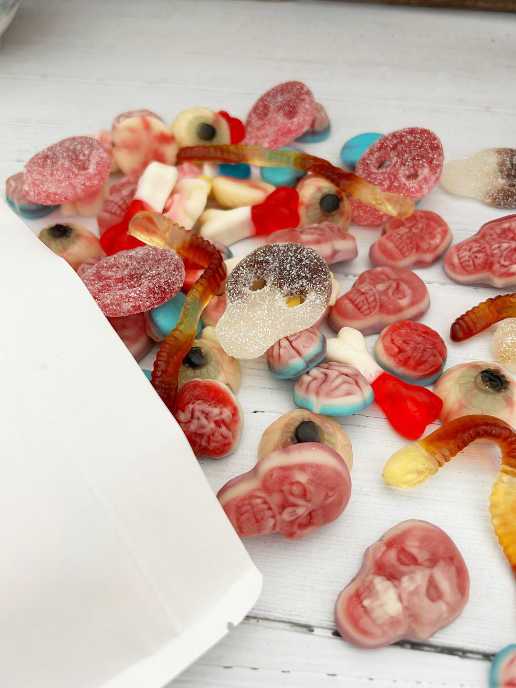 A close-up of assorted pick 'n' mix sweets spilled onto a white wooden surface, including brain-shaped gummies, worm gummies, and eye gummies.