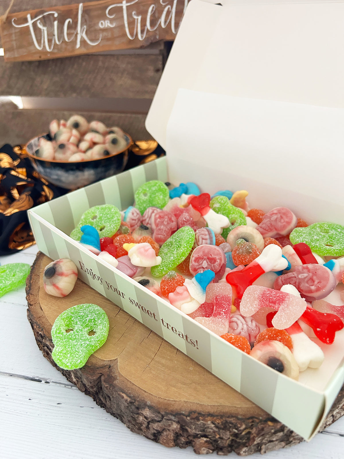 A box of assorted Halloween gummy candies, fizzy skulls, jelly eyeballs etc all displayed on a rustic wood slice with a bowl of gummy eyeballs and a 'Trick or Treat' sign in the background.