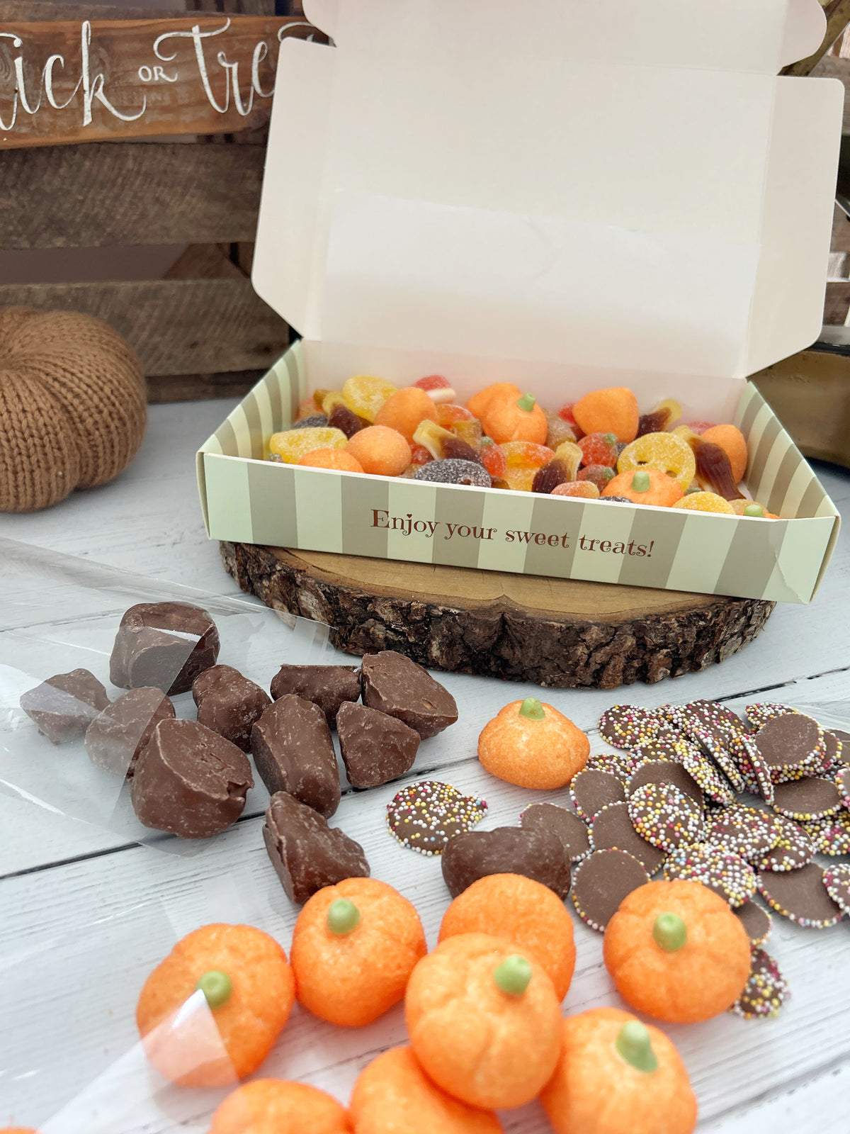 An open striped gift box filled with assorted Halloween treats, including orange pumpkin-shaped sweets, chocolate pieces, and sprinkle-coated jazzies, displayed on a rustic wood slice with more treats arranged around it.
