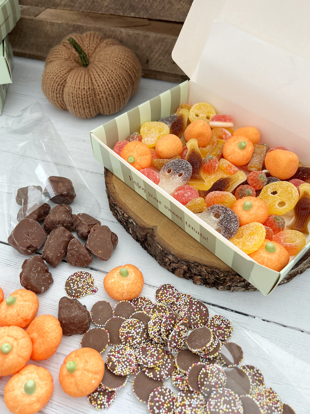 A box of assorted Autumn treats and sweets displayed on a rustic wood slice, with orange pumpkin-shaped sweets, chocolate pieces, and sprinkled jazzies arranged around it on a white wood surface.