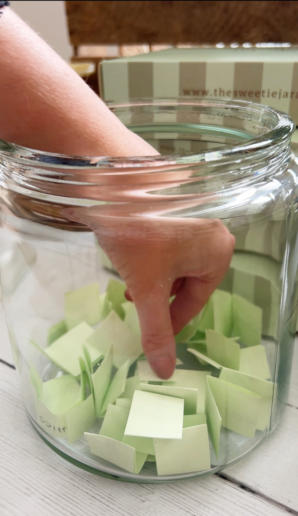 A hand placing light green sticky notes into a large clear glass jar on a white wooden surface.
