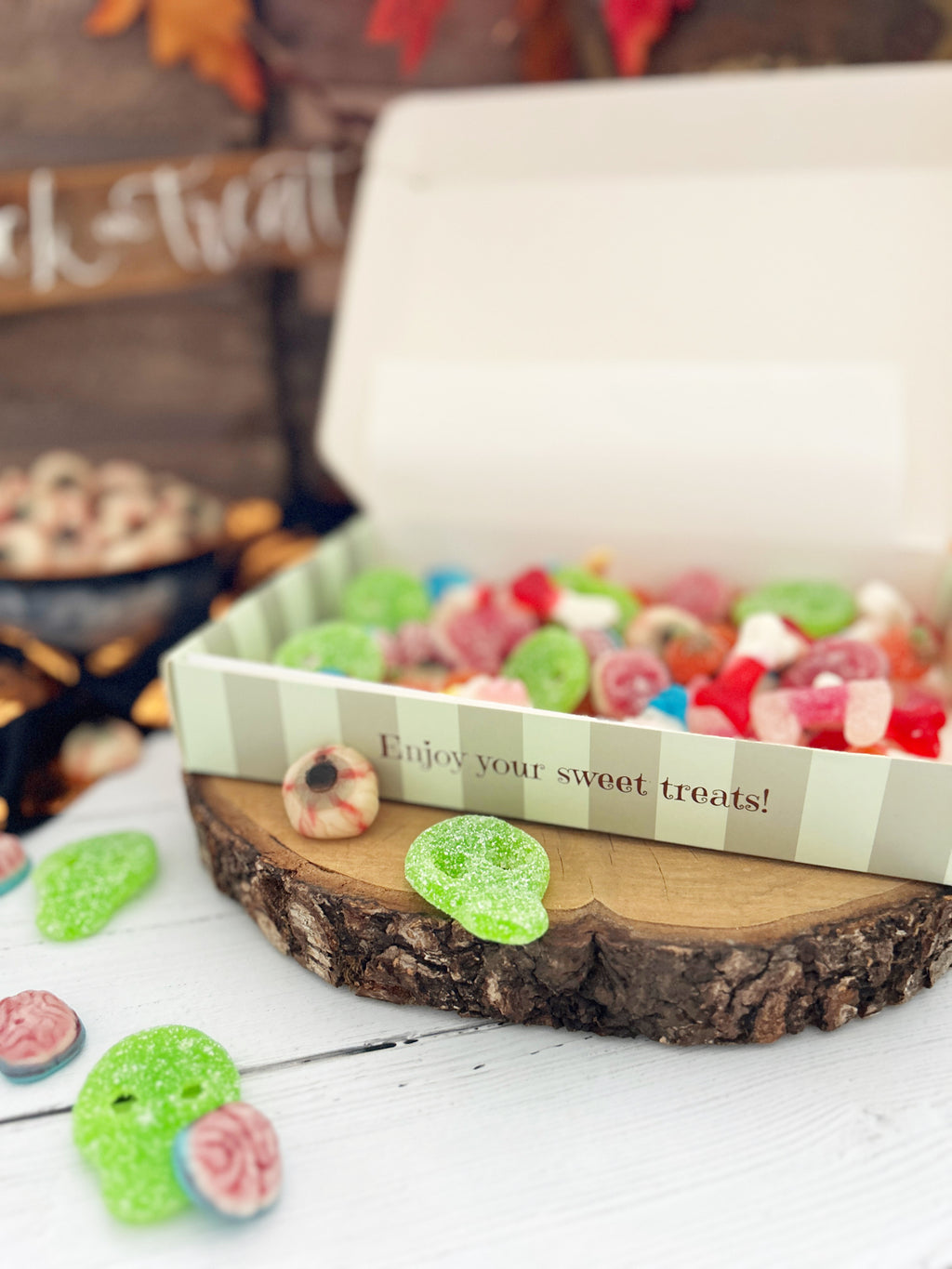 A box of colorful gummy candies displayed on a rustic wood slice stand, with a few candies scattered nearby and a Halloween-themed blurred background.