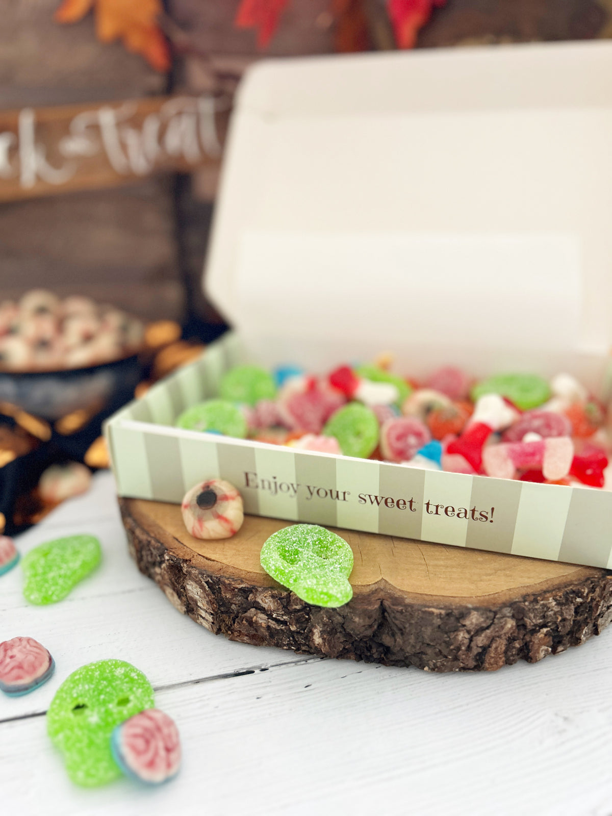 A box of colorful gummy candies displayed on a rustic wood slice stand, with a few candies scattered nearby and a Halloween-themed blurred background.