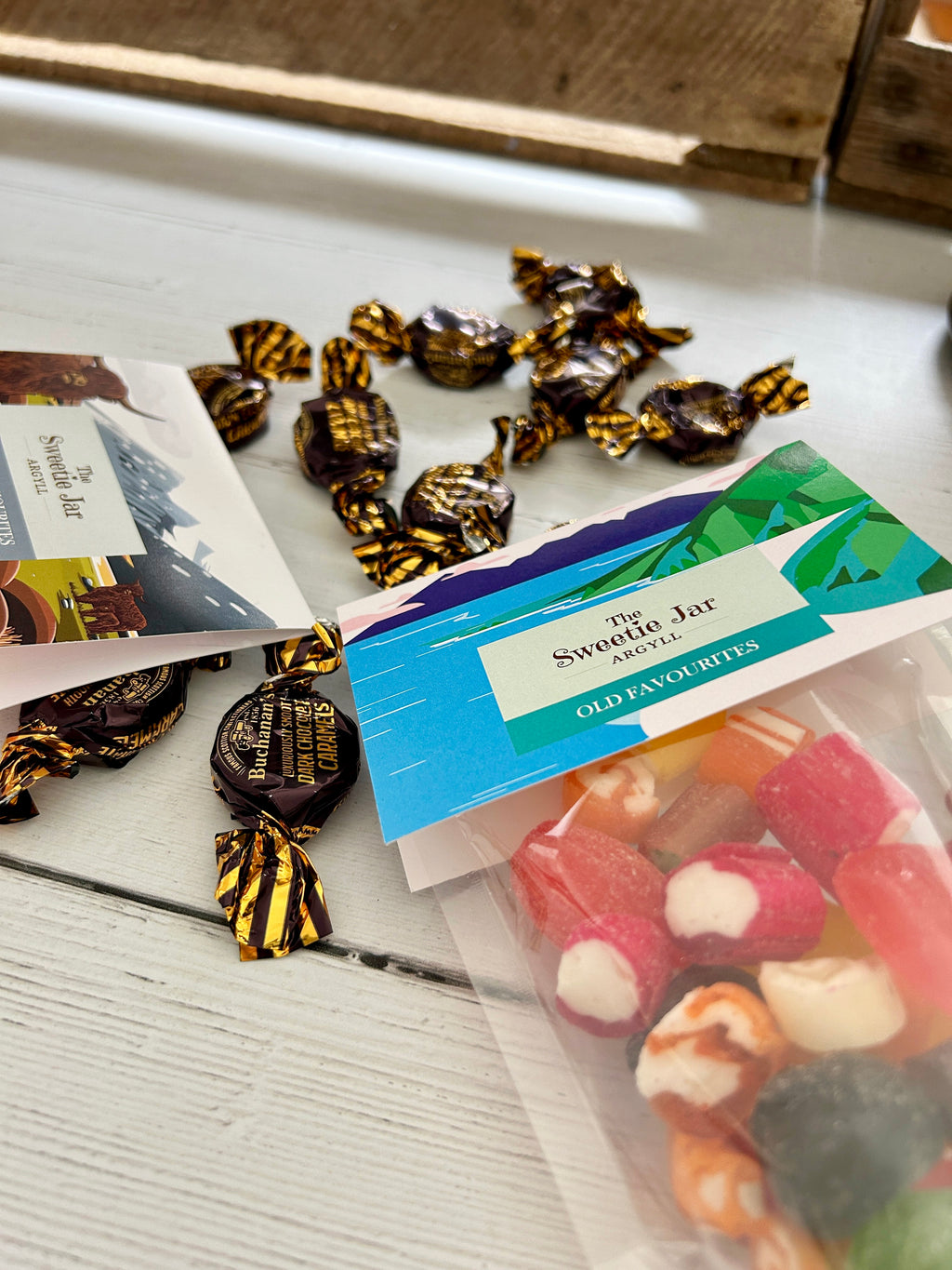 Two clear bags of Sweetie Jar sweets and chocolates arranged on a white wooden surface, with several individually wrapped dark chocolates scattered around them. One bag contains bright pink/red boiled candies and the other contains assorted colourful candies.