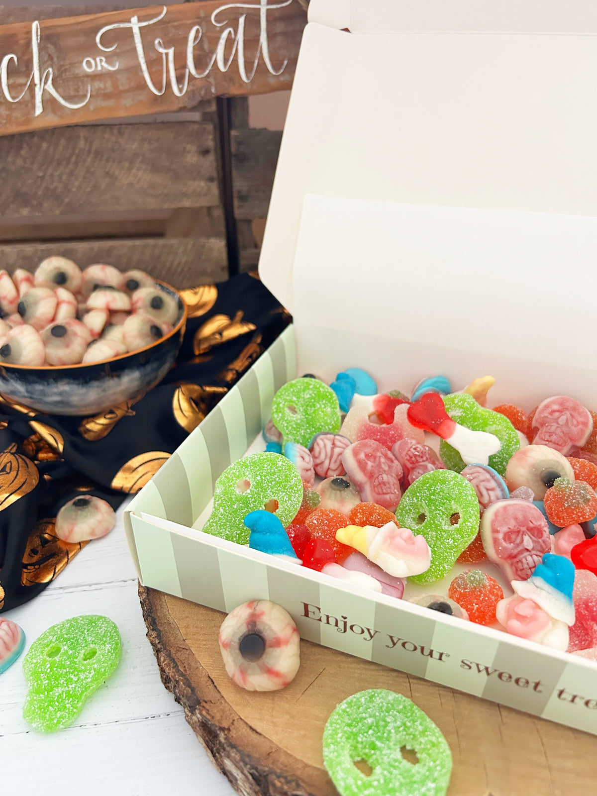 A box of colorful Halloween-themed gummy candies, including green gummy skulls, ghost gummies, and eyeballs, displayed on a rustic wood slice with a bowl of eyeballs gummies in the background.