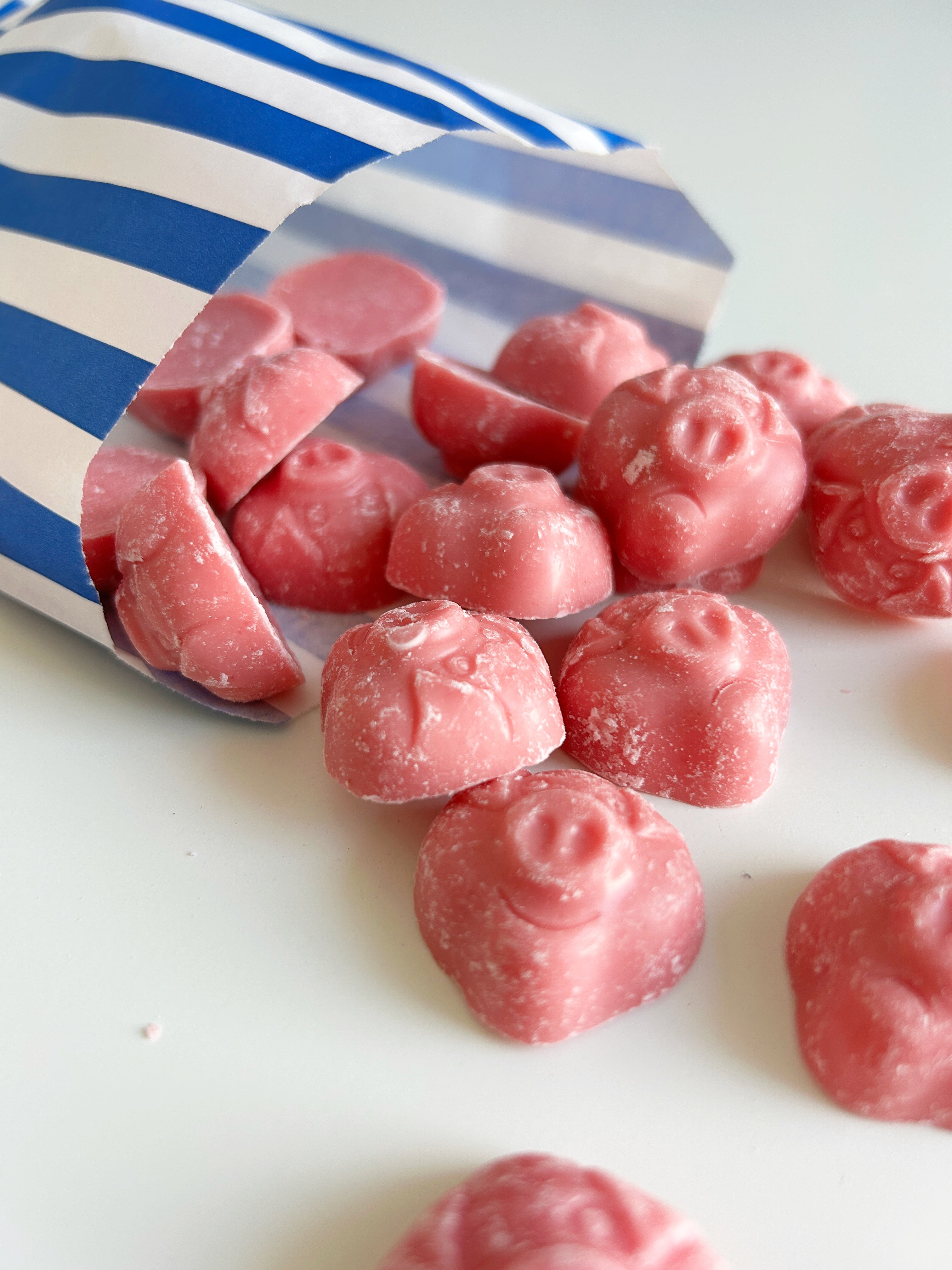 Pink, pig shaped candies spilling out of a blue and white striped paper bag onto a white surface.