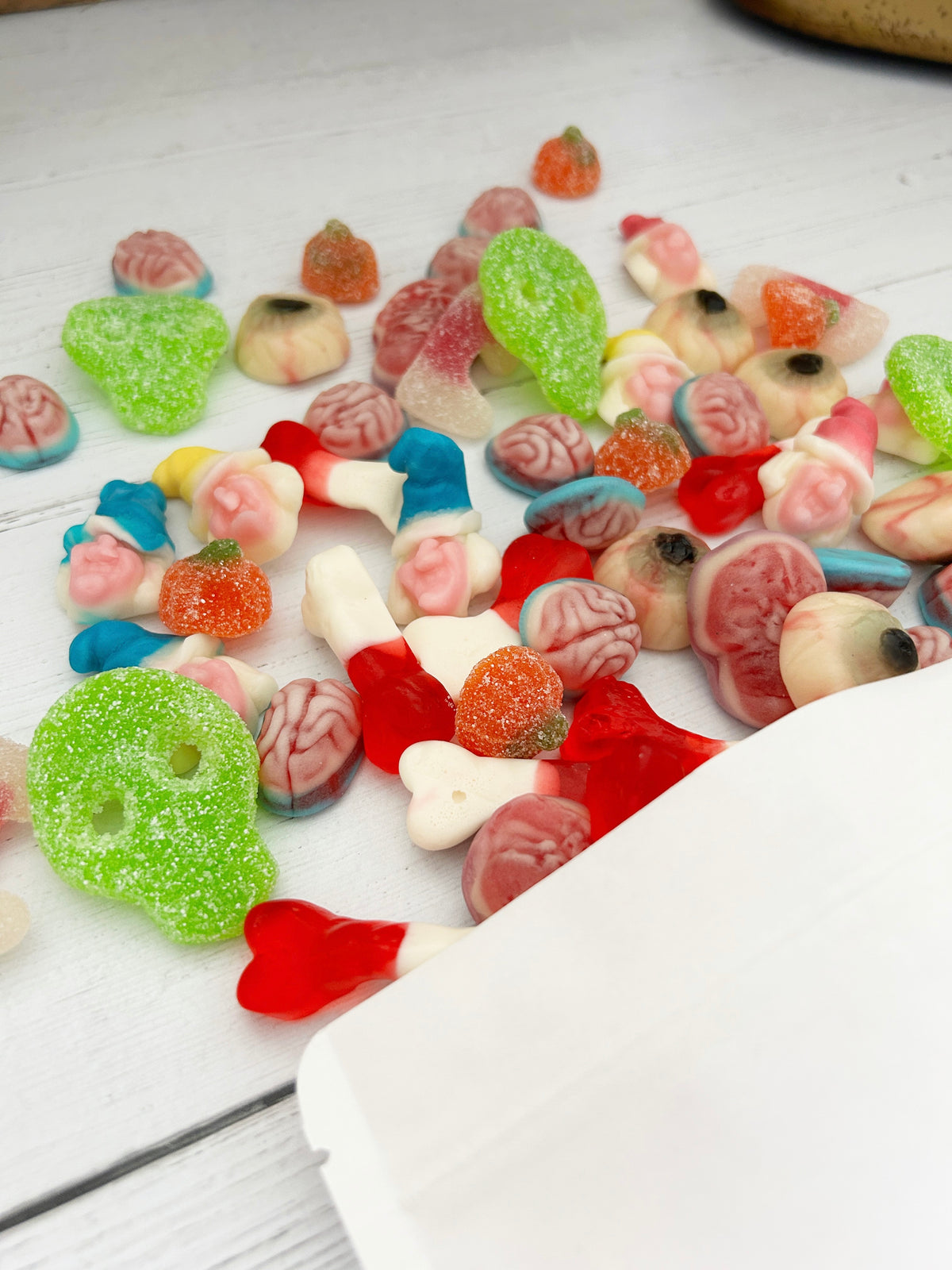 A variety of colorful Halloween-themed gummy candies spilled onto a white wooden surface, including brain-shaped gummies, skull gummies, vampire fangs, and pumpkin gummies, with a white box corner visible in the foreground.