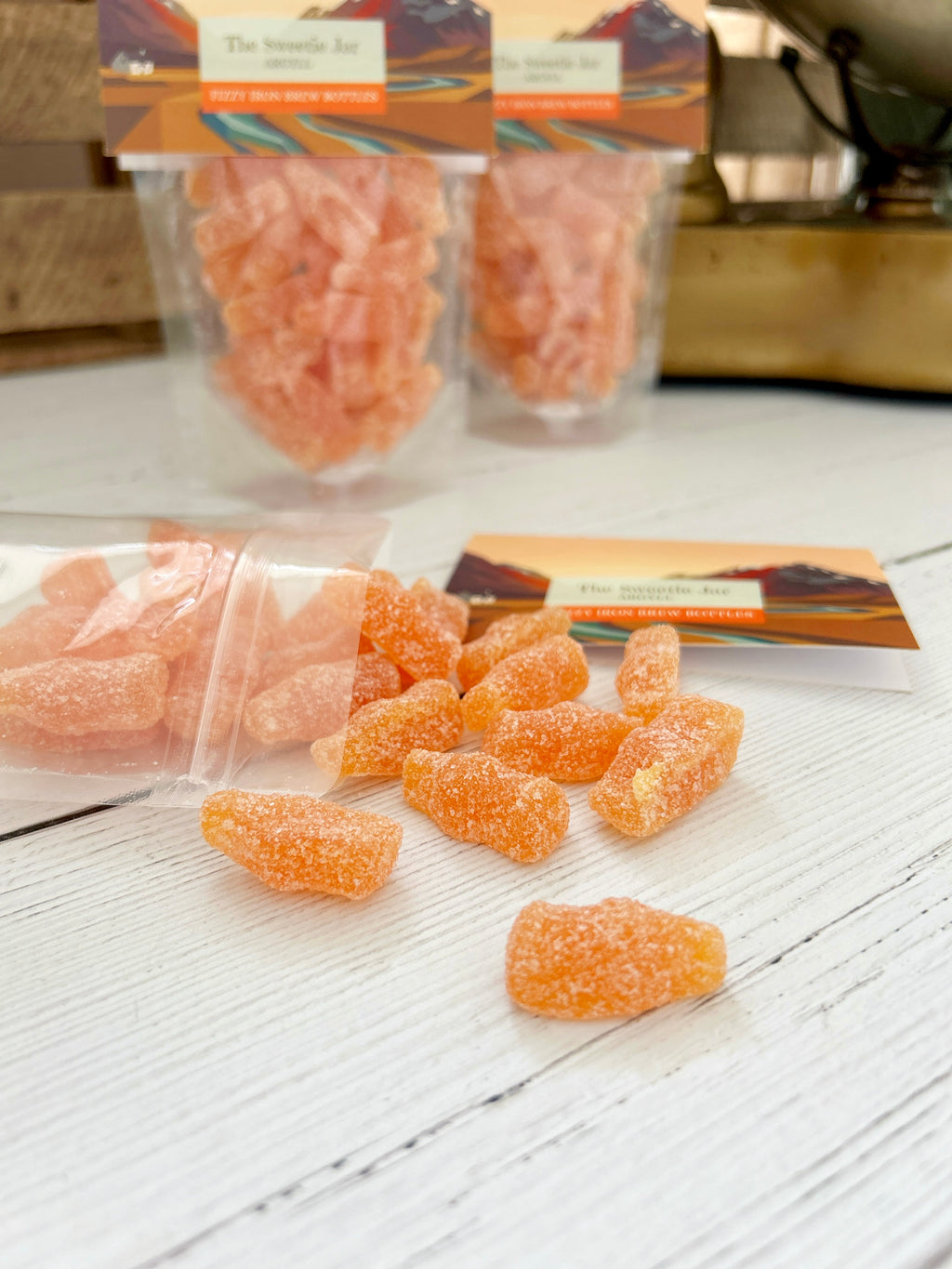 Orange fizzy iron brew bottles scattered on a white wooden surface, with two clear bags of the same candy and one loose bag in the background. The packaging has a warm multicolor label.