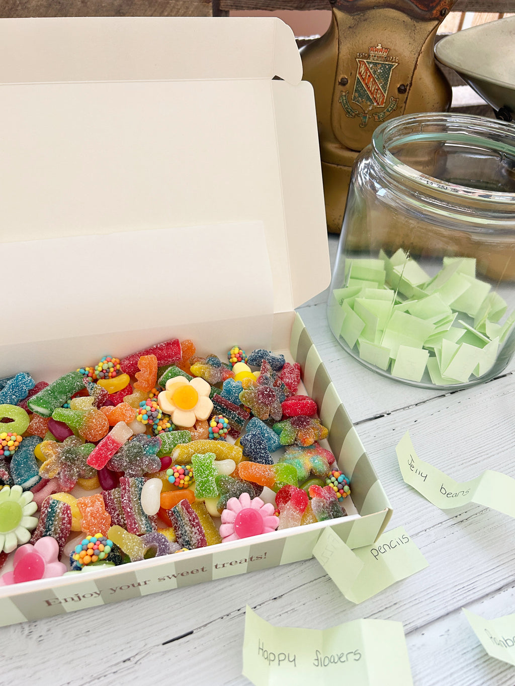 A white gift box filled with assorted colorful gummy candies and sweets sits open on a white wooden table, next to a glass jar of green candy pieces and small handwritten paper notes.