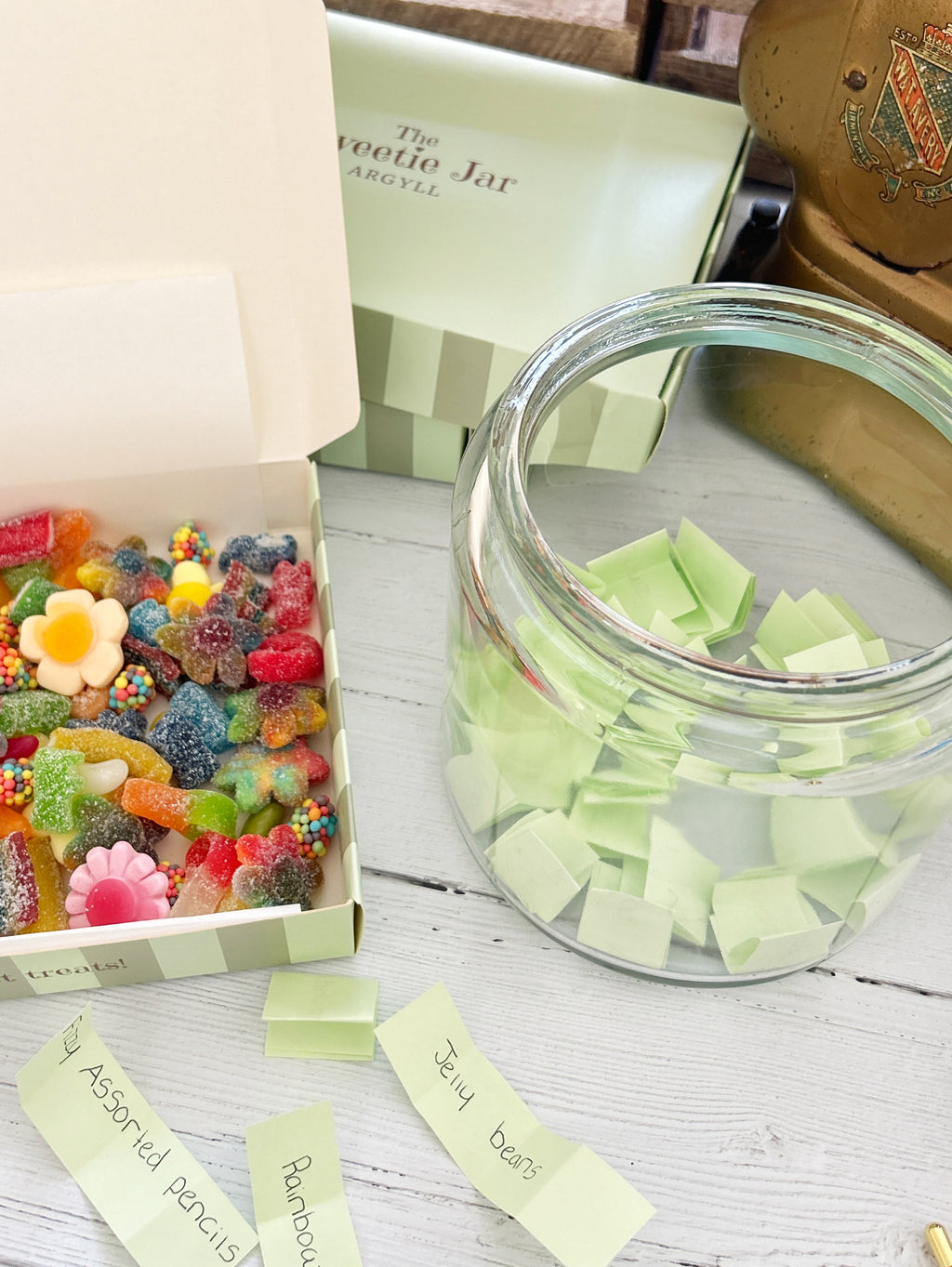 A themed candy gift set featuring a clear glass jar filled with pale green sticky notes, an open box of assorted colorful gummy candies, and handwritten name tags on a white wooden table. The brand 'The Sweetie Jar' is visible on the packaging.