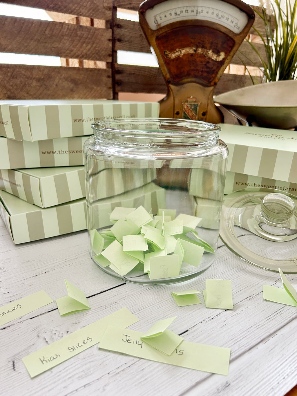 A clear glass jar filled with pale green folded paper notes sits on a white wooden surface, with matching striped boxes and a vintage-style balance scale in the background.