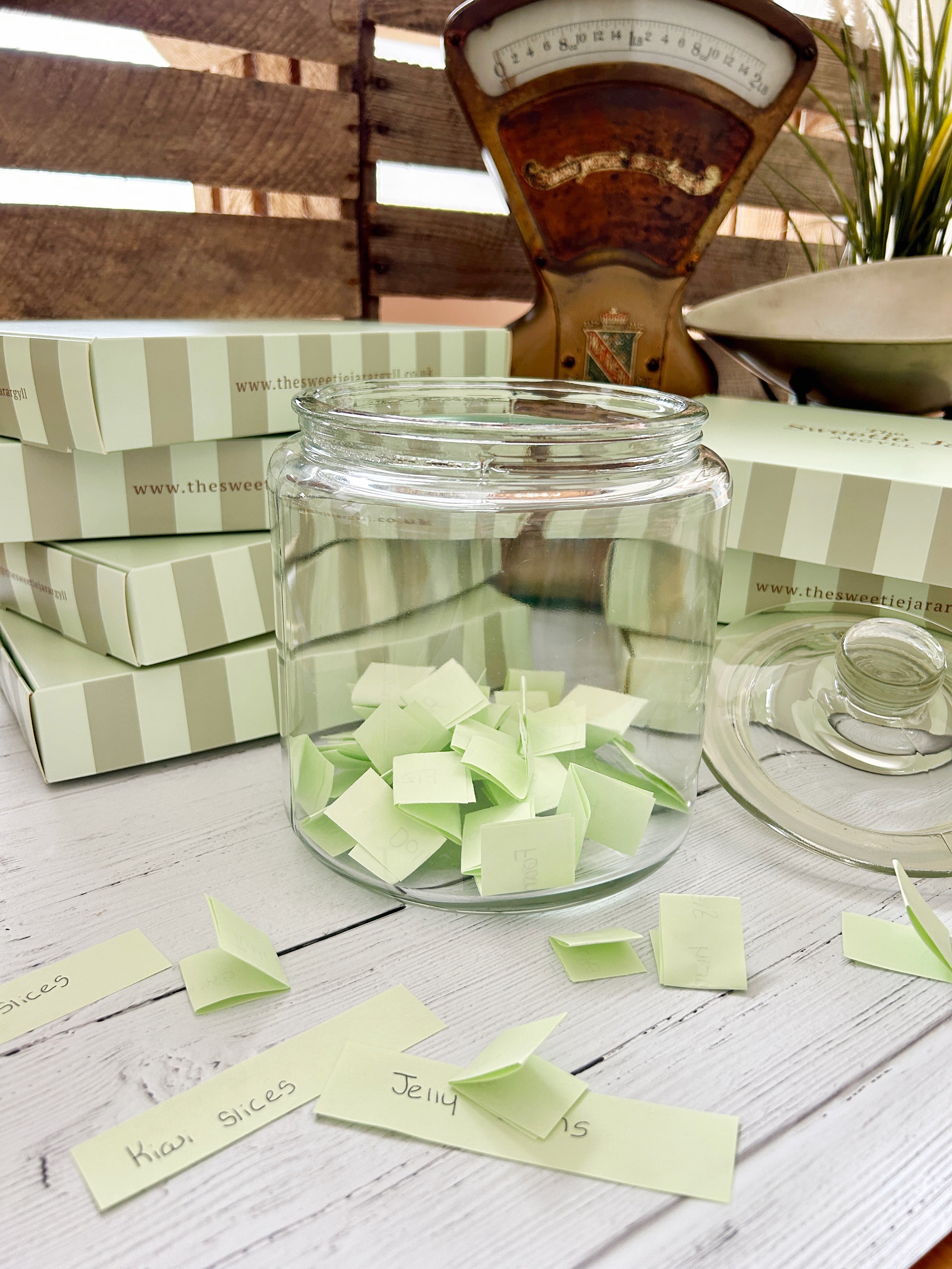 A clear glass jar filled with pale green folded paper notes sits on a white wooden surface, with matching striped boxes and a vintage-style balance scale in the background.