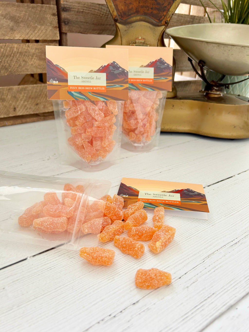 Two clear stand-up pouches of orange iron brew bottles with printed labels are displayed on a white wooden surface, with some candy spilled in front. A vintage brass scale and rustic wood background are visible behind the product.